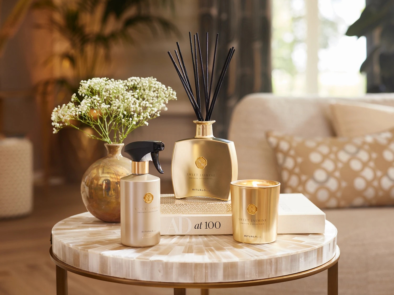 Auksinės spalvos „Rituals“ namų kvapų rinkinys su jazminų aromatu, žvakė ir purškiklis ant staliuko su gėlėmis, sukuriantis jaukią atmosferą.