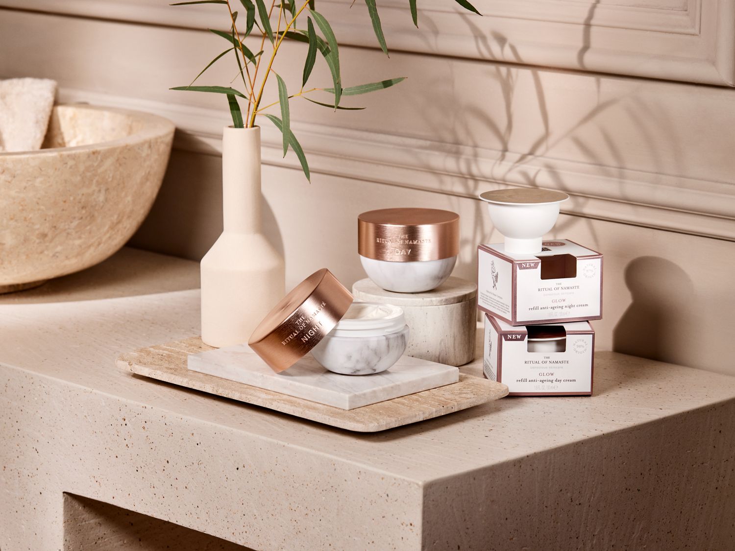„Ritual of Namaste Glow“ kolekcija: marmuriniai indai su rožinio aukso dangteliais dieniniam ir naktiniam kremui, papildomi kremai dėžutėse, vazos su augalais ant smėlio spalvos paviršiaus.