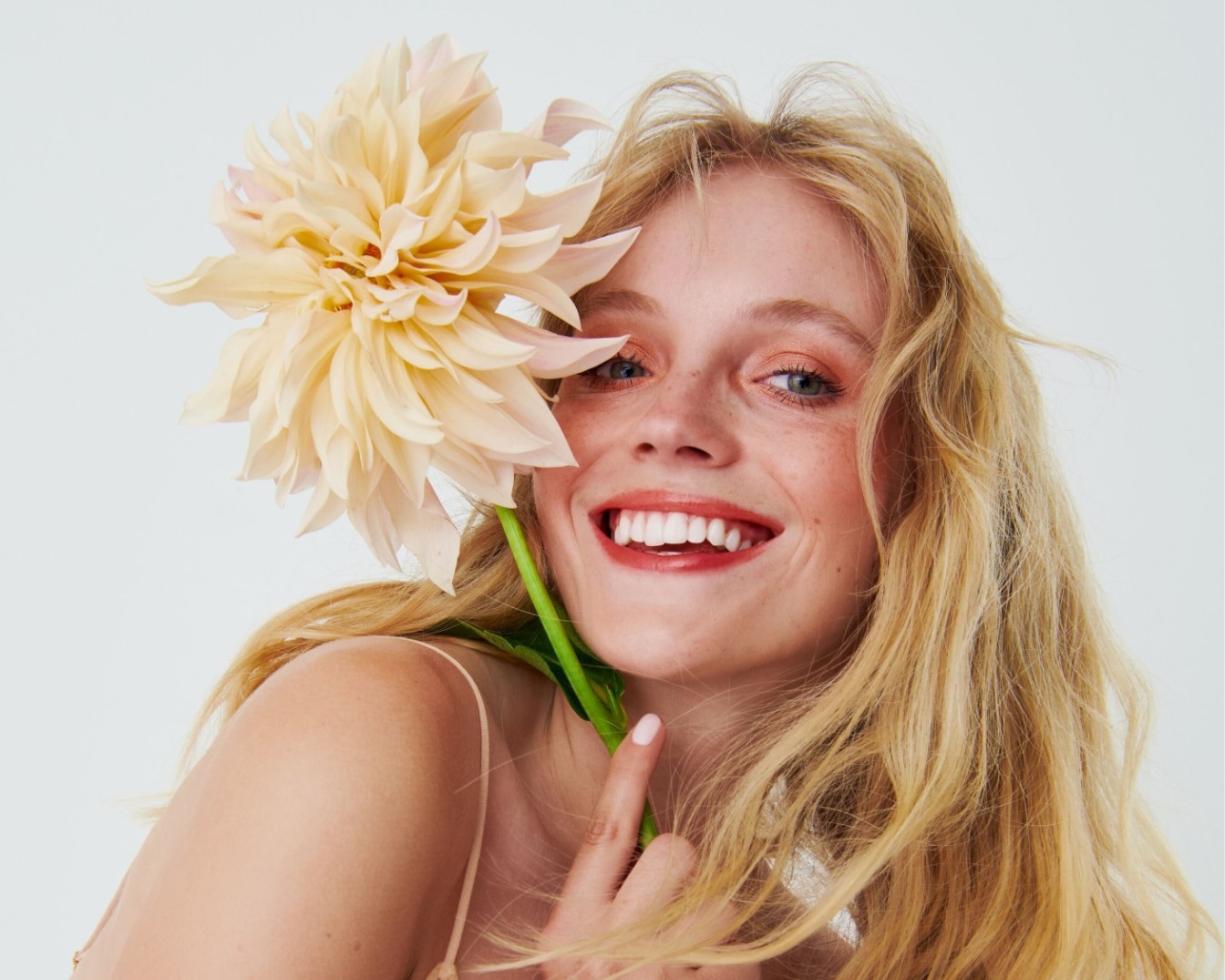 Usmievavá žena s blond vlasmi drží žltú kvetinu pri tvári, jej úsmev zvýrazňuje rúž.