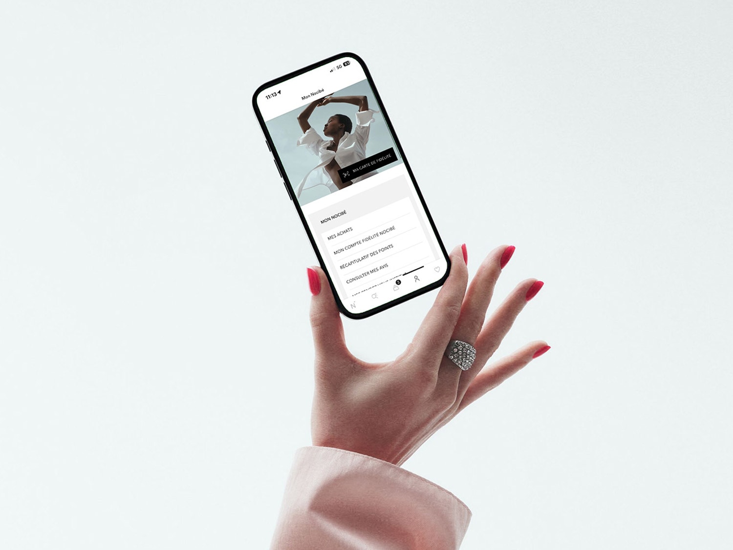 Découvrez l'application Mon Nocibé sur un smartphone tenu par une main élégante avec un vernis rouge éclatant et une bague étincelante, offrant un accès facile à votre compte, vos achats et vos points de fidélité.