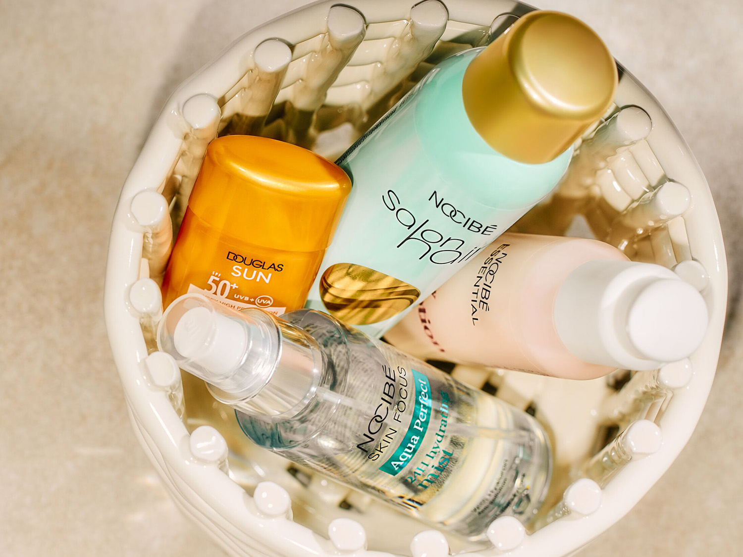 Dans un panier en céramique, découvrez une sélection de produits de beauté : un écran solaire Douglas SPF 50, un soin capillaire Nocibé, une lotion essentielle Nocibé et une brume hydratante Aqua Perfect de Nocibé.
