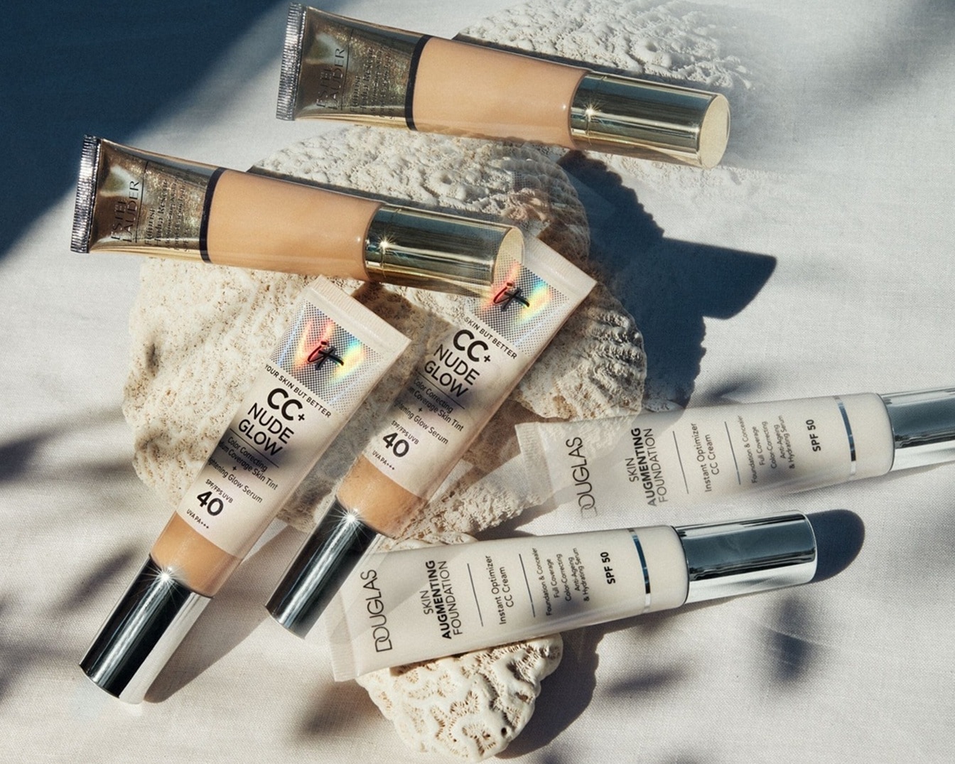 Verschillende foundations en CC-crèmes van merken als Estée Lauder, IT Cosmetics en Douglas liggen uitgestald op een lichte achtergrond.