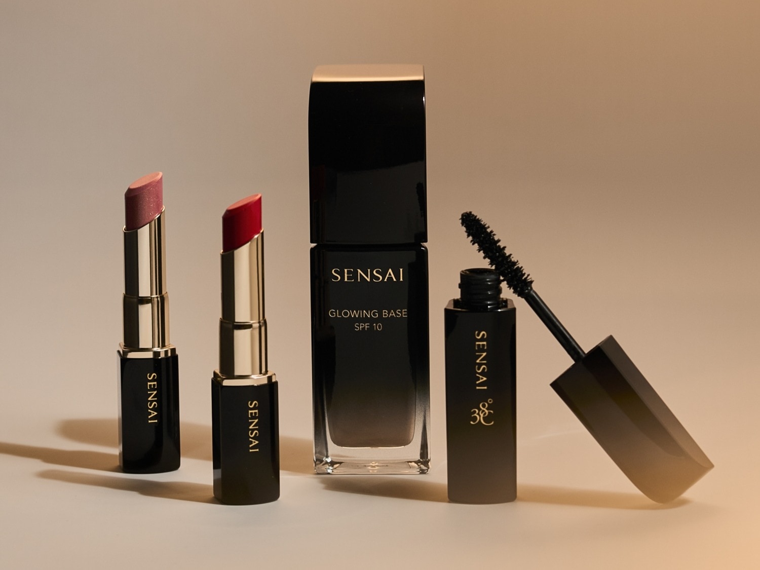 Découvrez un assortiment de produits de beauté Sensai, comprenant deux rouges à lèvres aux teintes rose et rouge, une base de teint lumineuse avec SPF 10, et un mascara 38°C pour un regard intense.