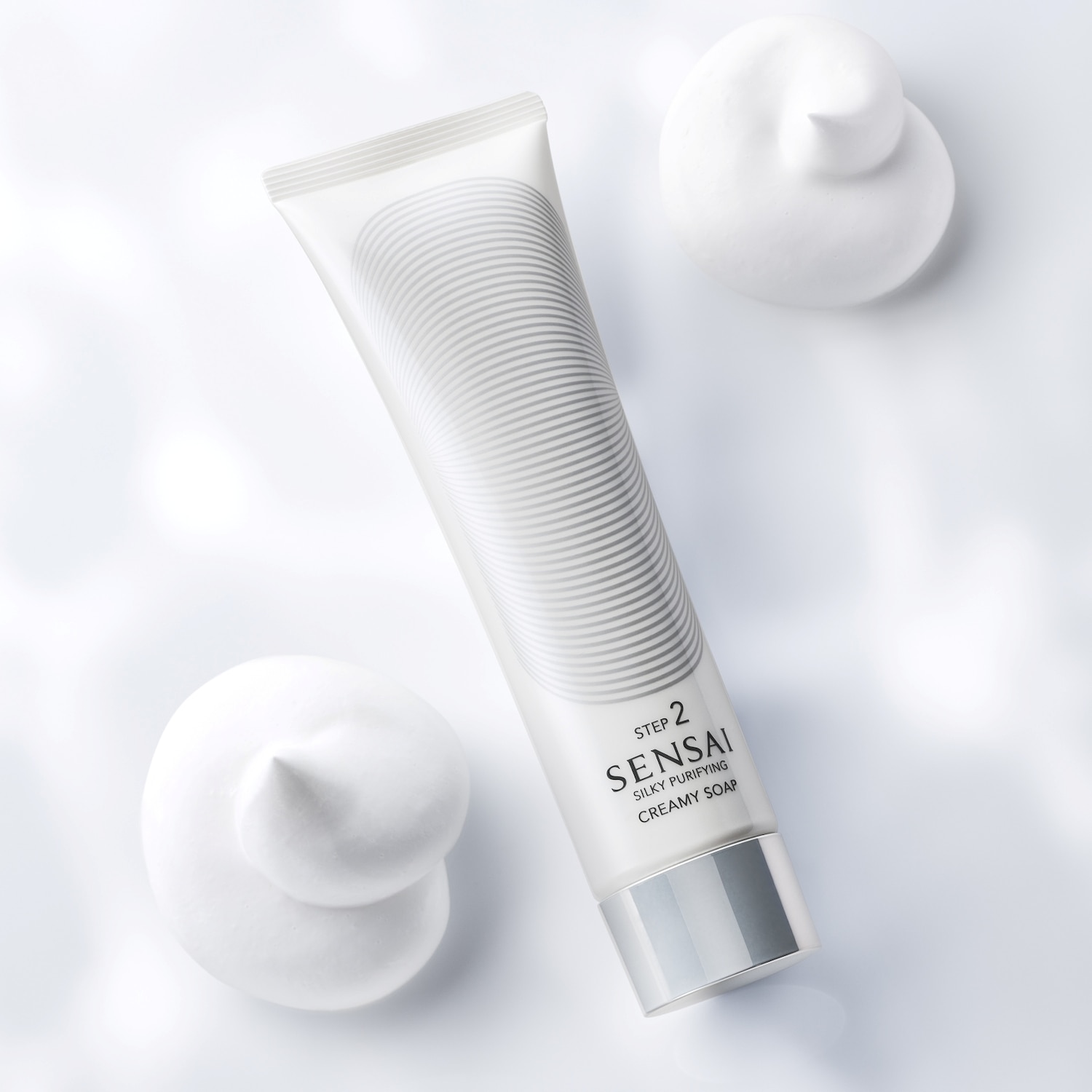 Découvrez le Savon Crème Purifiant Soyeux Sensai, étape 2, dans son tube blanc orné de lignes argentées, accompagné de touches de mousse crémeuse.