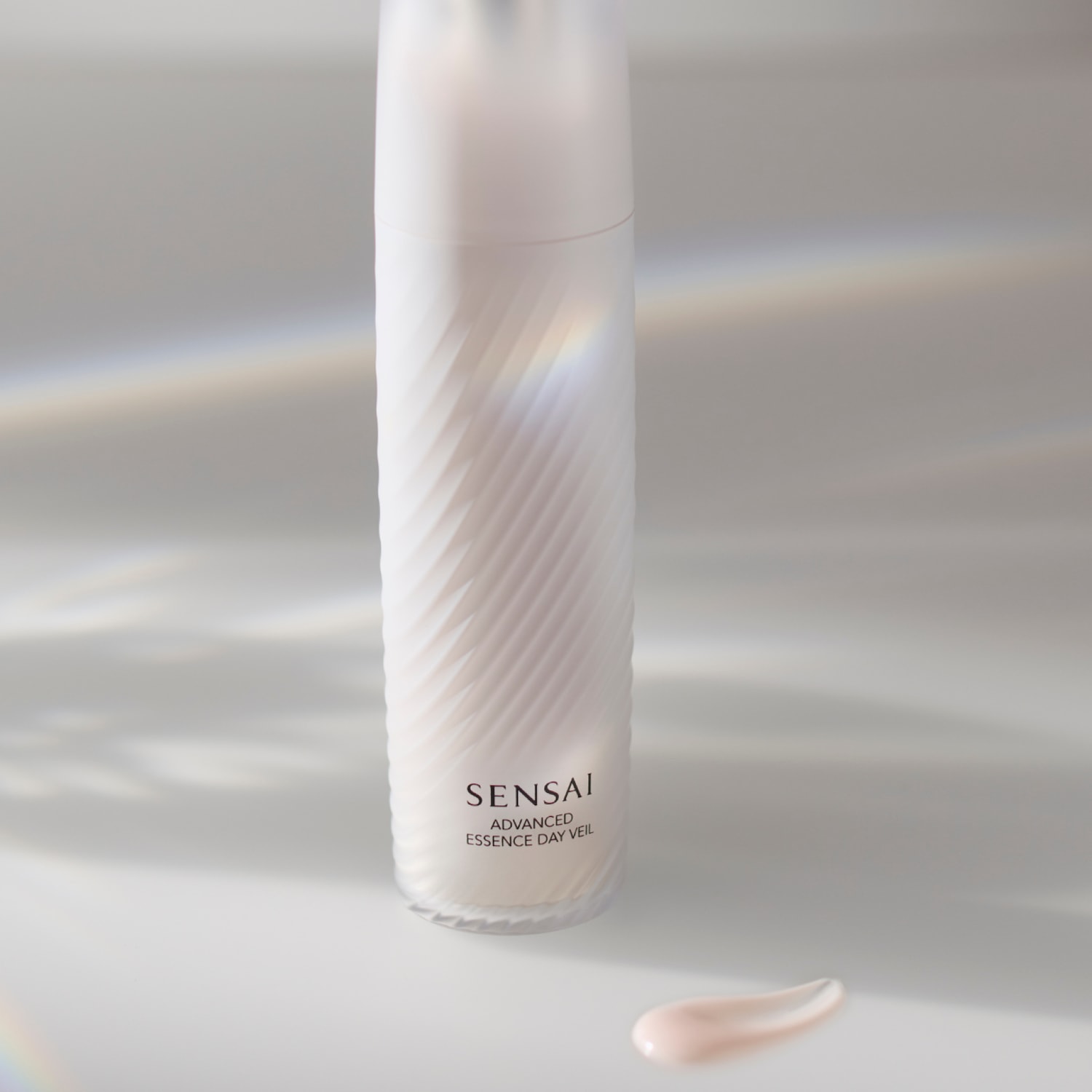 Présentation du voile de jour essence avancée SENSAI dans un flacon blanc texturé, avec une touche de crème rose pâle à côté.