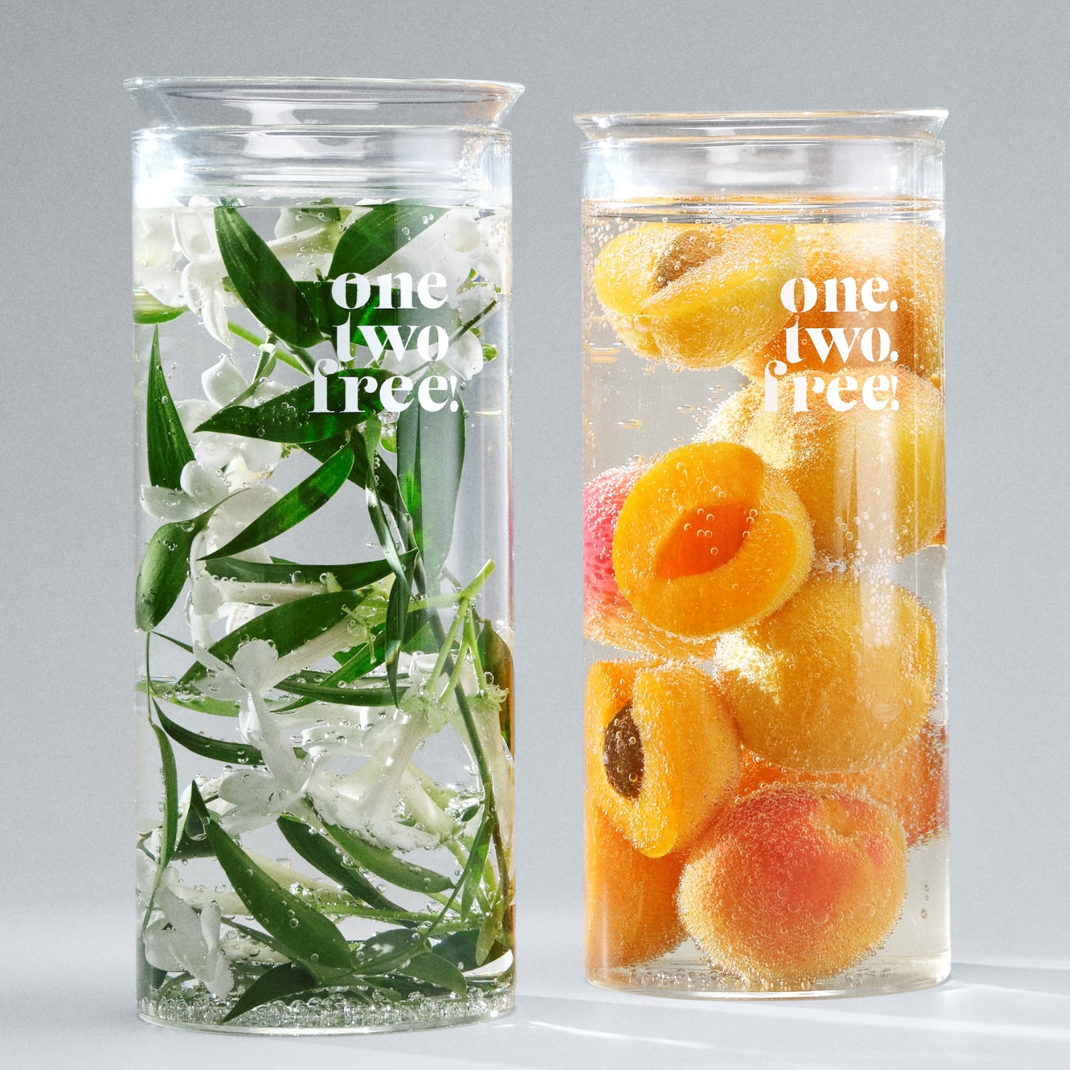 Zwei Glasbehälter, einer mit weißen Blüten und grünen Blättern in Wasser, der andere mit Aprikosen und Pfirsichen in Wasser, beide tragen den Schriftzug "one two free!".