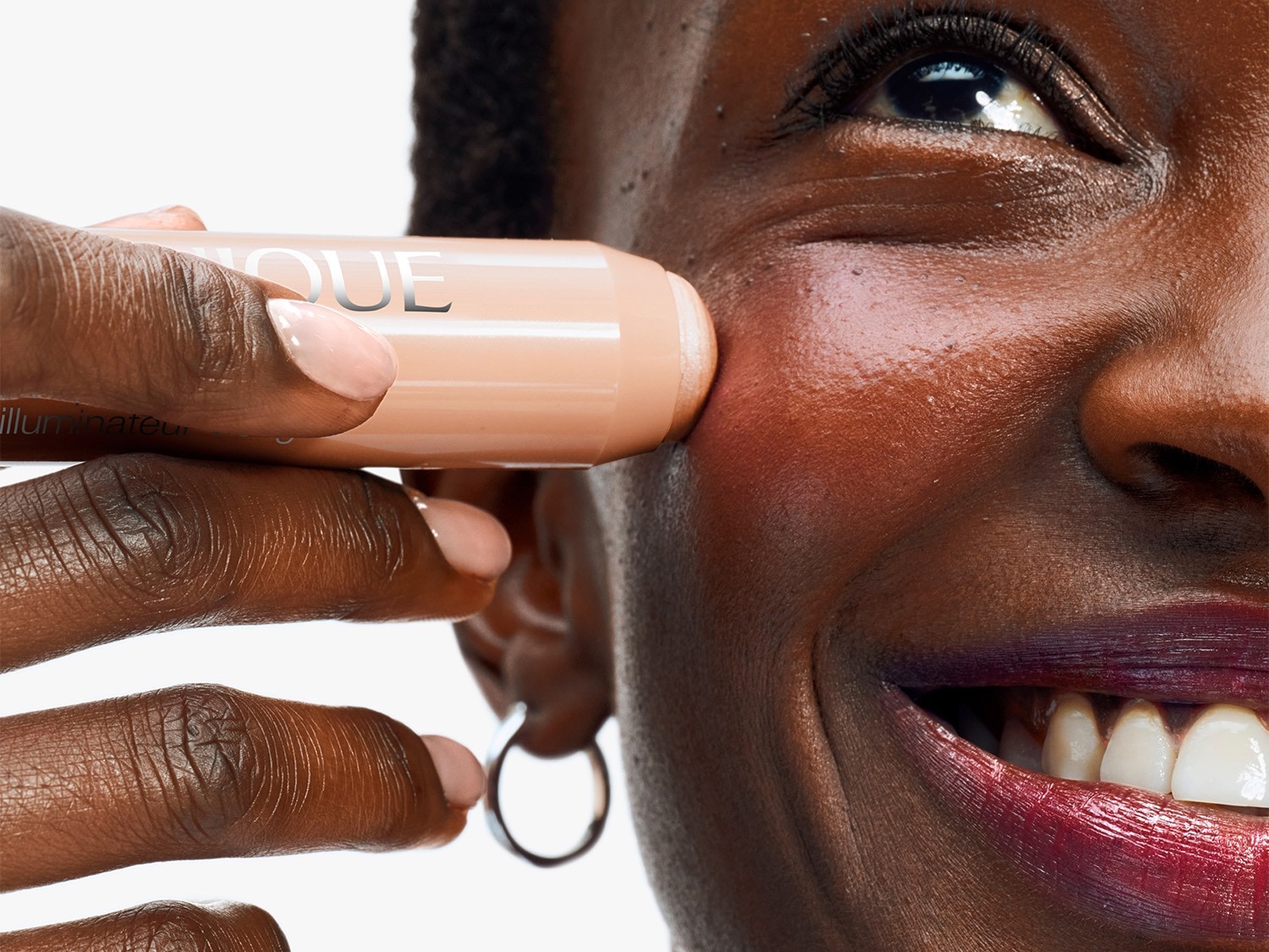 Een vrouw met een stralende glimlach brengt een Clinique highlighter aan op haar wang, voor een subtiele, verhelderende gloed.