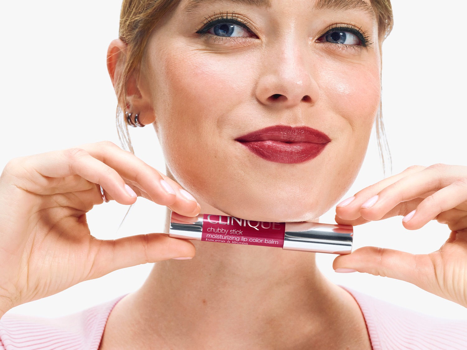Vrouw met blauwe ogen en roze trui houdt Clinique Chubby Stick Moisturizing Lip Color Balm vast, lippenstift is bessenkleurig.