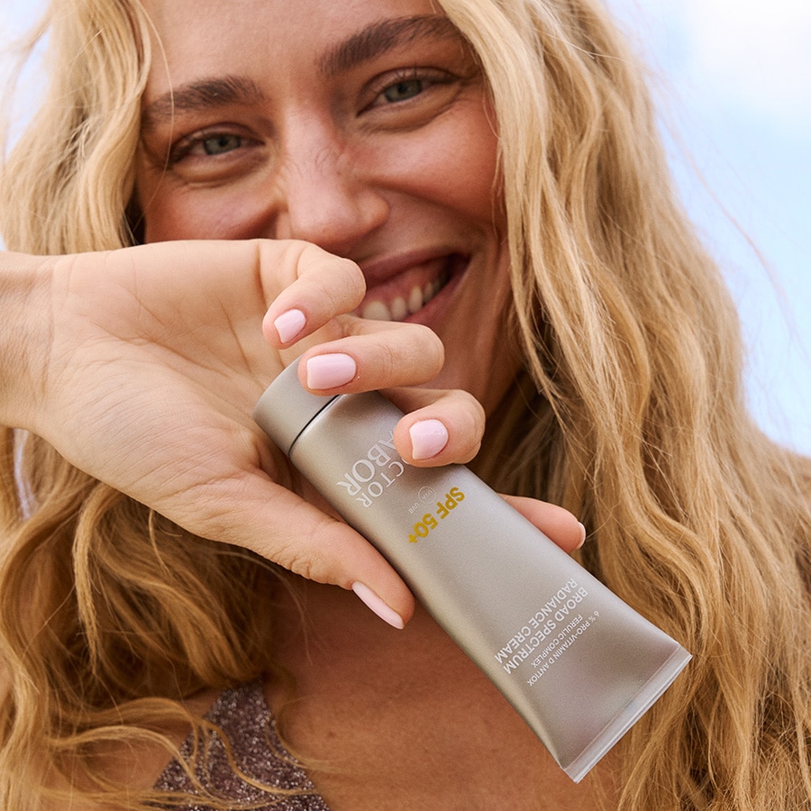 Eine strahlende Frau hält eine Tube Babor Broad Spectrum Radiance Cream SPF 50+ in der Hand, die 6 % Pro-Vitamin D Antioxid Ferulic Complex enthält.