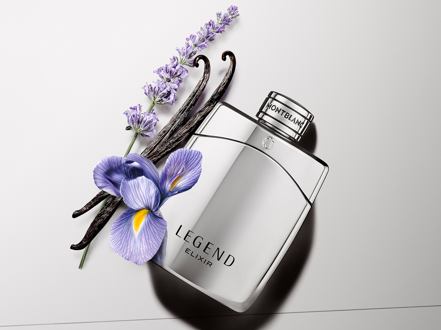 Srebrny flakon perfum Montblanc Legend Elixir otoczony kwiatami lawendy, irysa i laskami wanilii na białym tle.