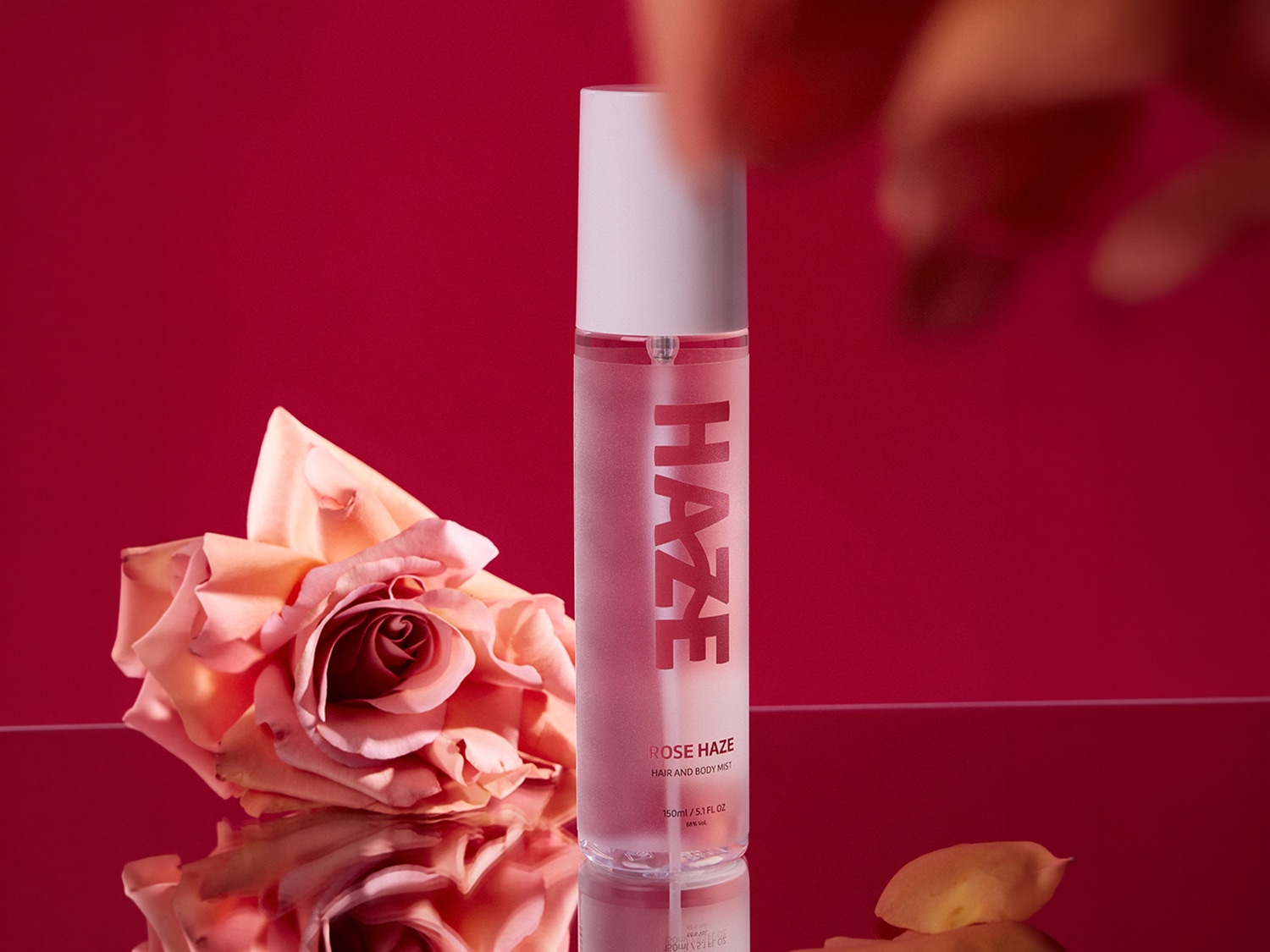Ecco una bottiglia di spray rosa per capelli e corpo Haze Rose, con un'etichetta bianca e una rosa color pesca accanto, su uno sfondo rosso.