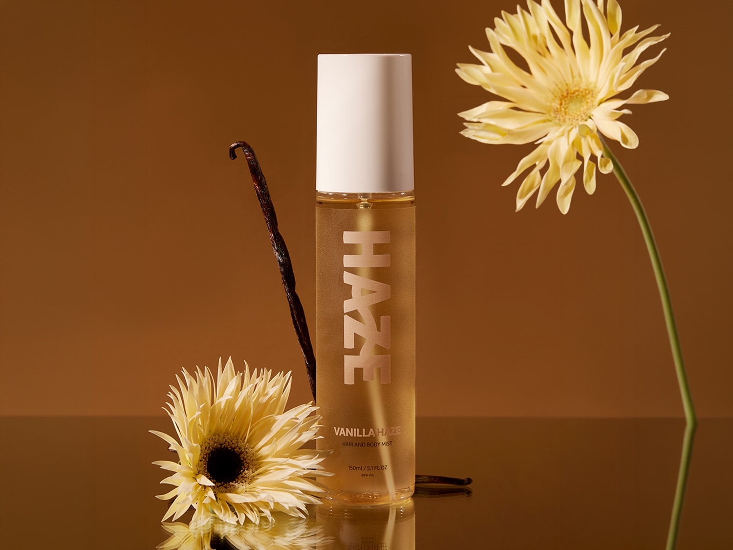 Ecco una bottiglia di spray per capelli e corpo Vanilla Haze, con un tappo bianco e un'etichetta dorata, adagiata accanto a fiori gialli e un baccello di vaniglia su uno sfondo marrone.