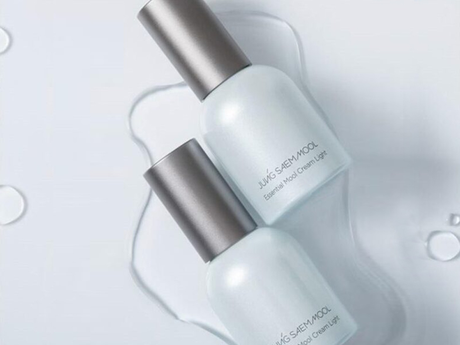 Dwa białe flakony kremu JUNG SAEM MOOL Essential Mool Cream Light z szarymi zakrętkami, otoczone kroplami wody, tworzą minimalistyczną i świeżą kompozycję.