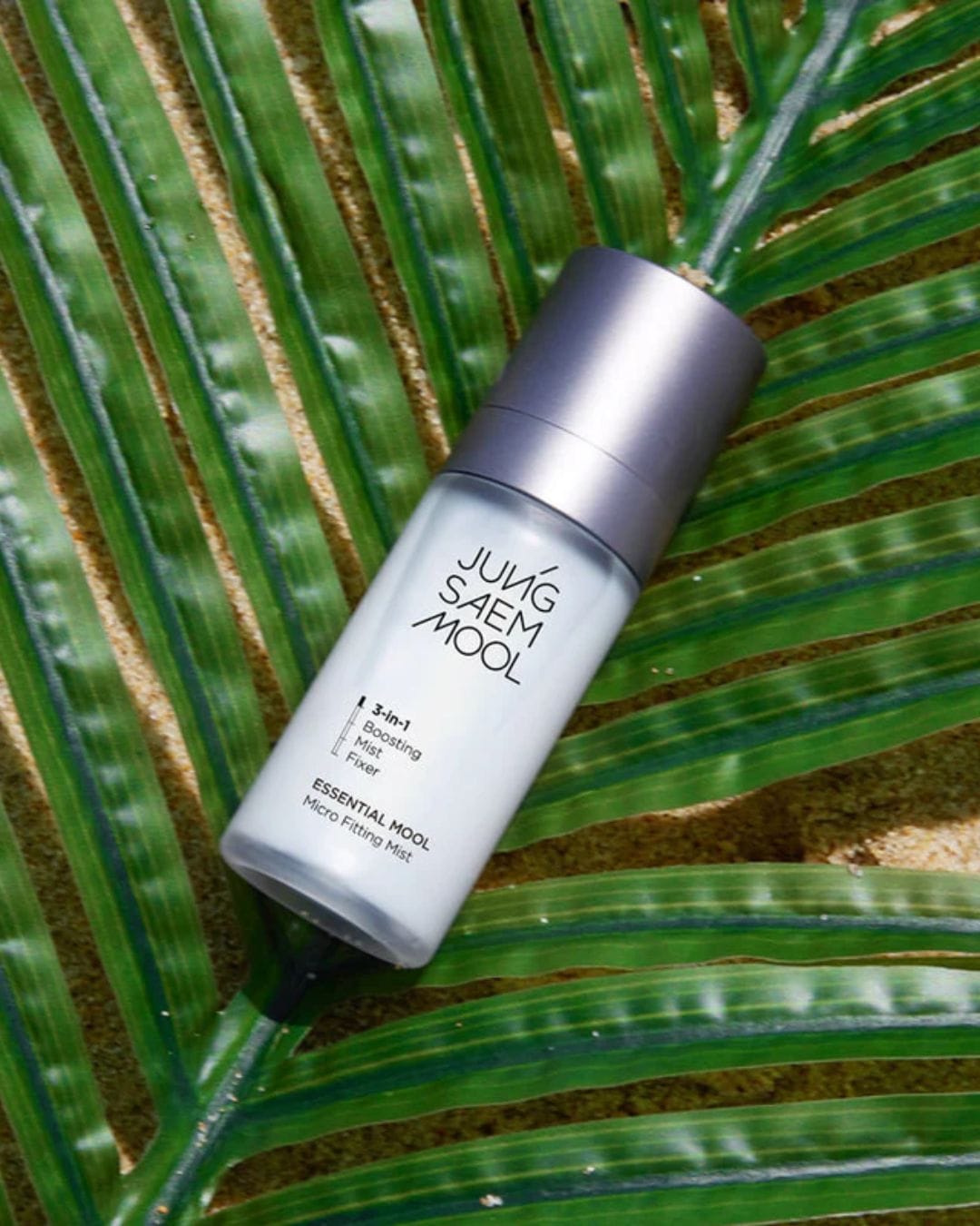 Biała butelka z rozpylaczem Jung Saem Mool Essential Mool Micro Fitting Mist 3 w 1 leży na zielonym liściu palmy.