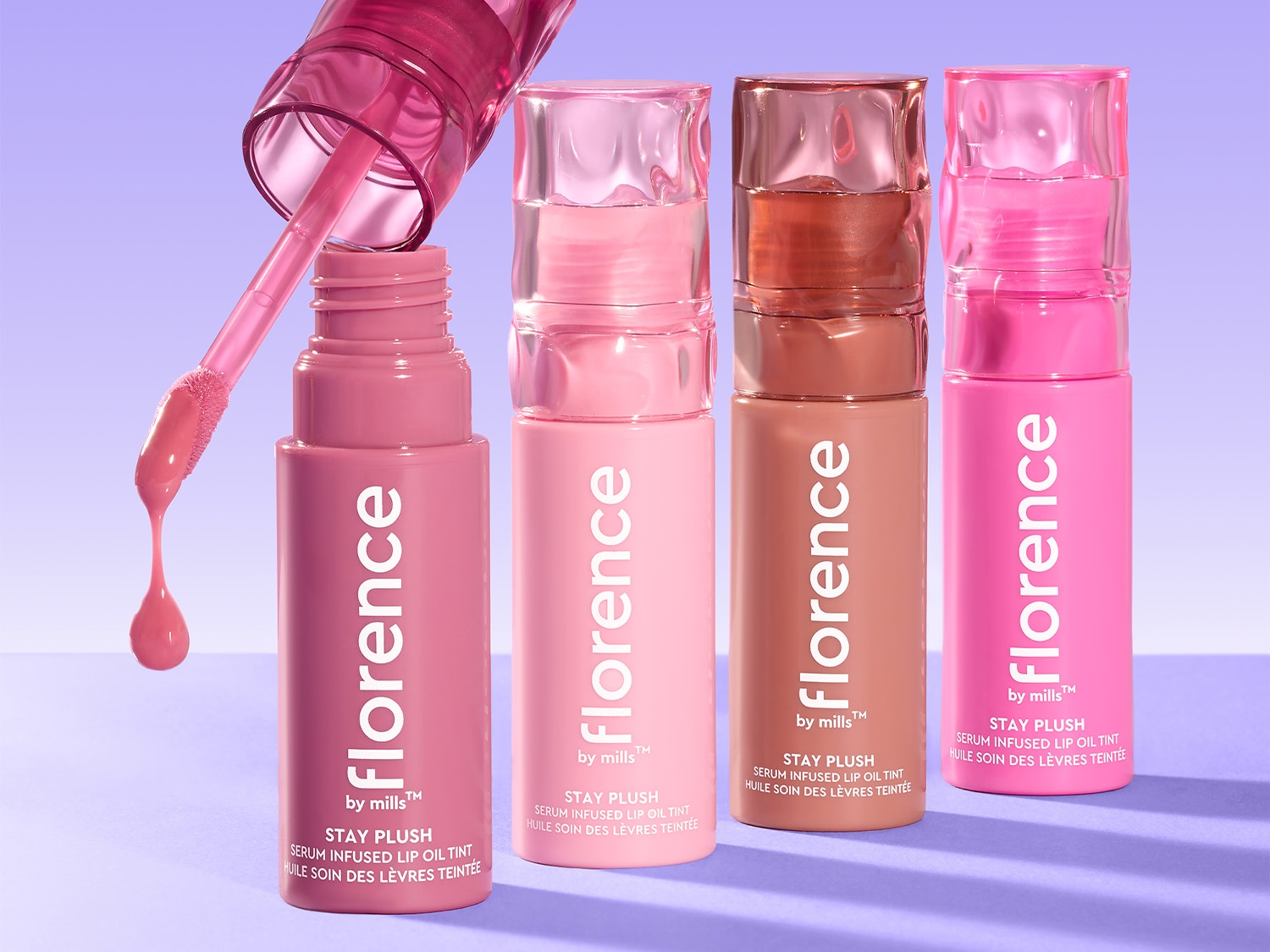 Keturių „Florence by Mills Stay Plush“ lūpų aliejų atspalvių kolekcija su skaidriais dangteliais ir aplikatoriumi, iš kurių vienas atidarytas, kad būtų parodyta produkto tekstūra.