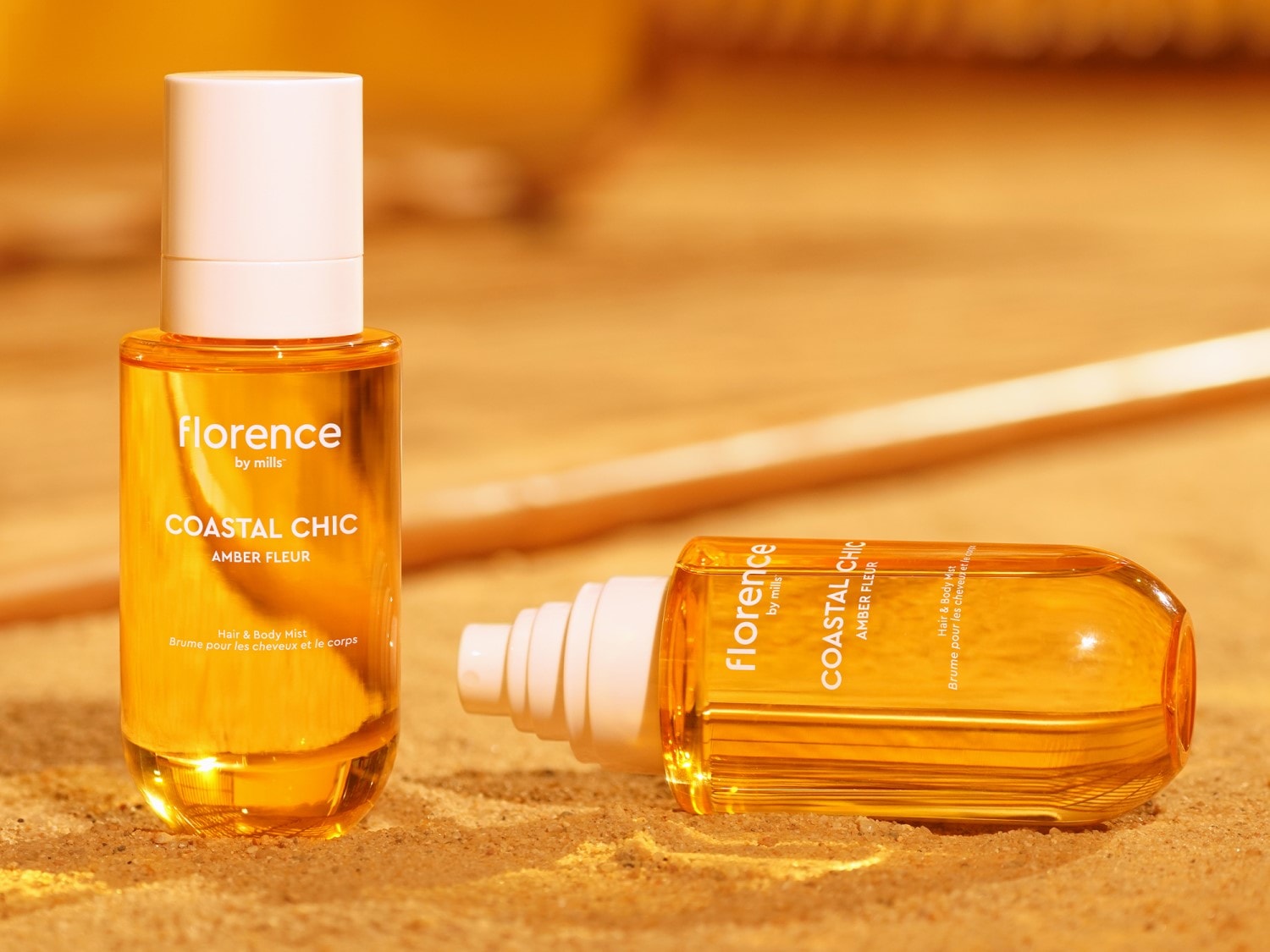 Du „Florence by Mills“ „Coastal Chic Amber Fleur“ plaukų ir kūno dulksnos buteliukai su baltu dangteliu, vienas stovi, kitas guli ant smėlio fone.