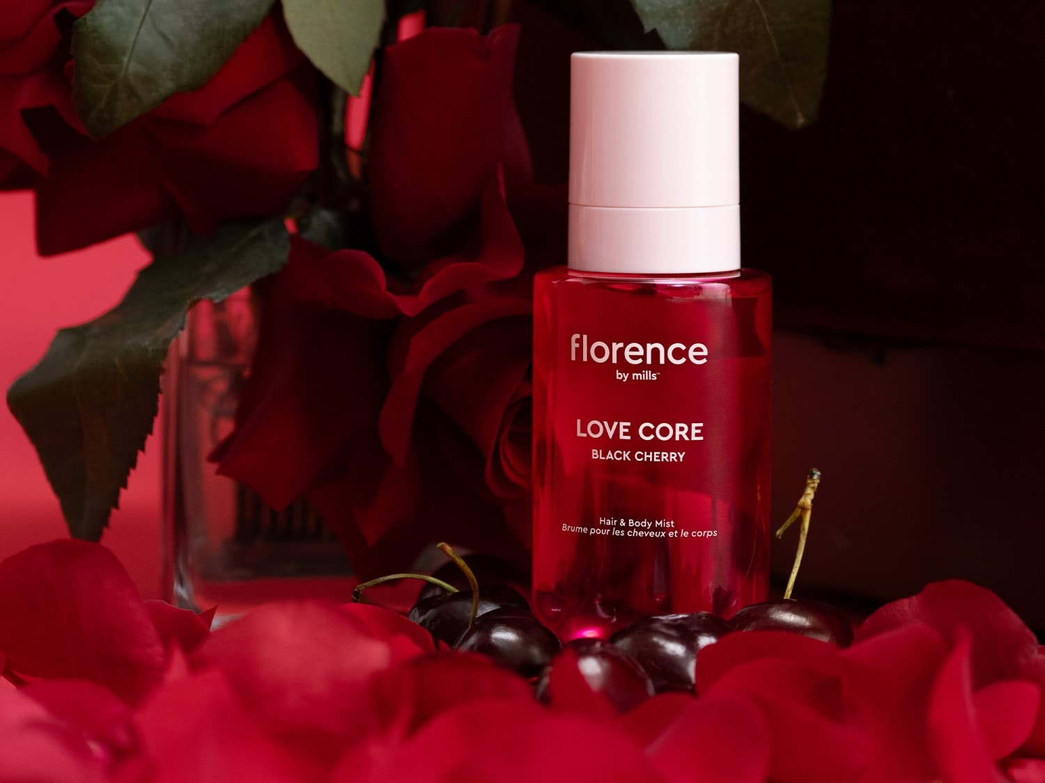 Florence by Mills Love Core Black Cherry plaukų ir kūno dulksna raudoname buteliuke su baltu dangteliu, apsupta raudonomis rožėmis ir vyšniomis.