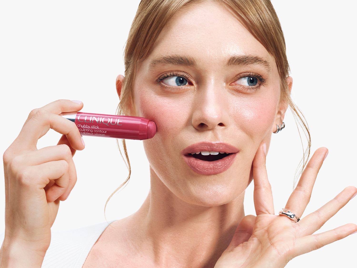 Une femme blonde applique un bâton de contour rose Clinique Chubby Stick sur sa joue, créant un look sculpté et rosé.