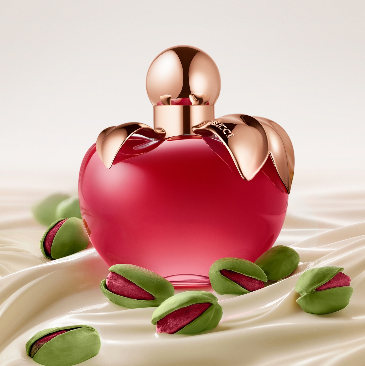 Découvrez Nina Ricci en flacon rouge en forme de pomme, orné de feuilles dorées, entouré de pistaches sur un tissu soyeux.