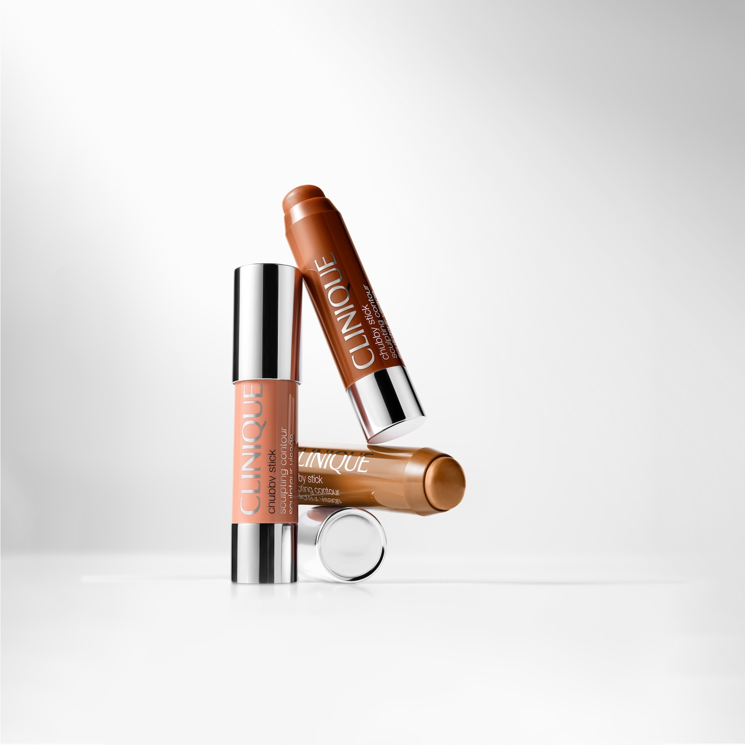 Eine Anordnung von drei Clinique Chubby Sticks Sculpting Contour in verschiedenen Farbtönen mit silbernen Deckeln, vor hellem Hintergrund.