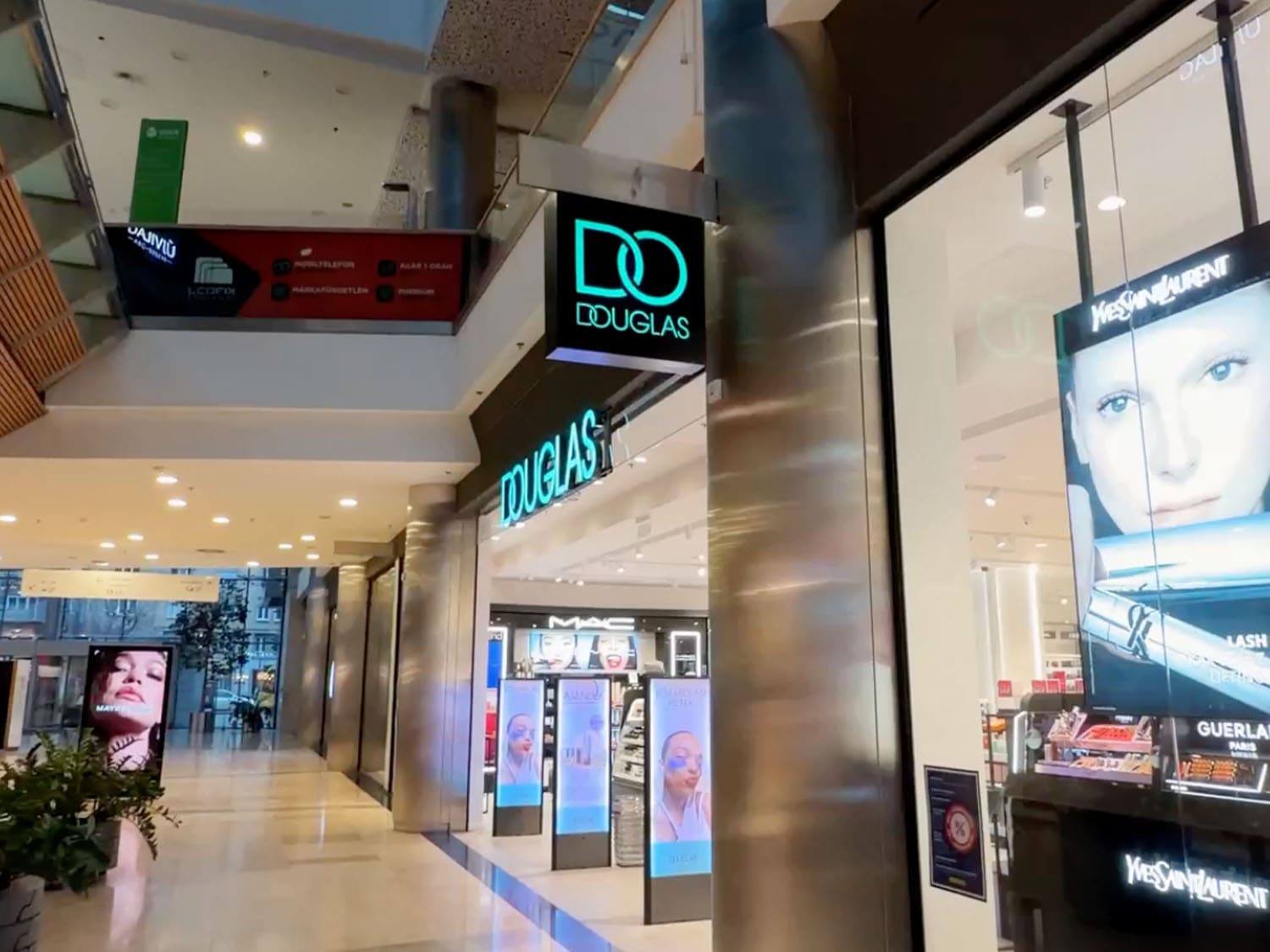 Pohled na obchod Douglas v nákupním centru s neonovým logem a reklamami značek Yves Saint Laurent a Guerlain ve výloze.