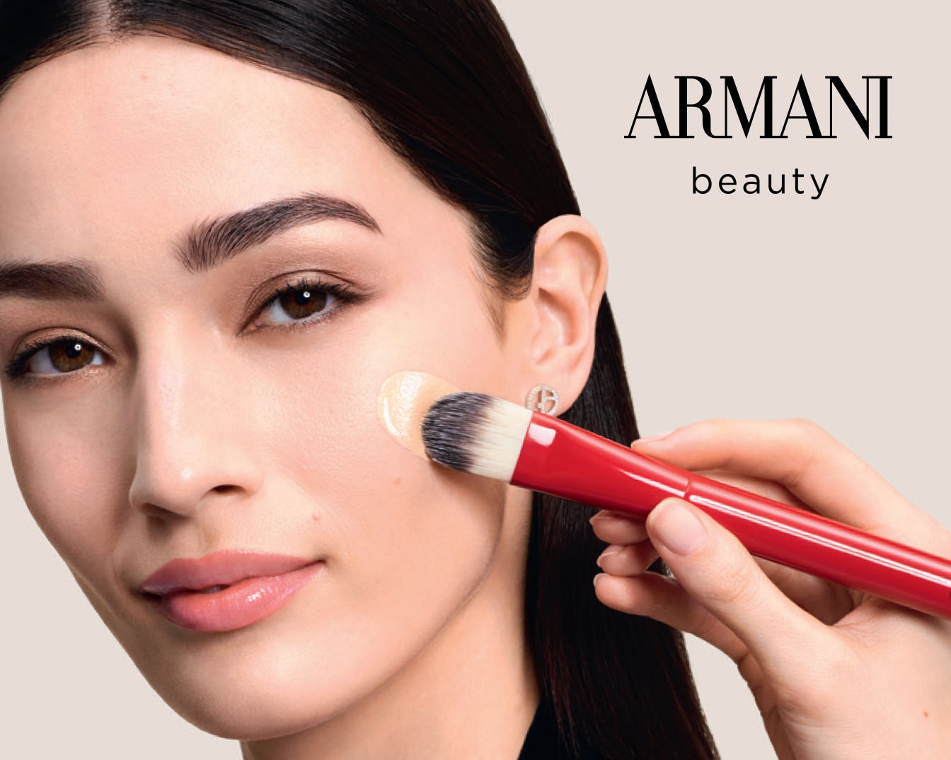Portrét ženy s hnědýma očima, která si štětcem s červenou rukojetí nanáší make-up na obličej od Armani Beauty.