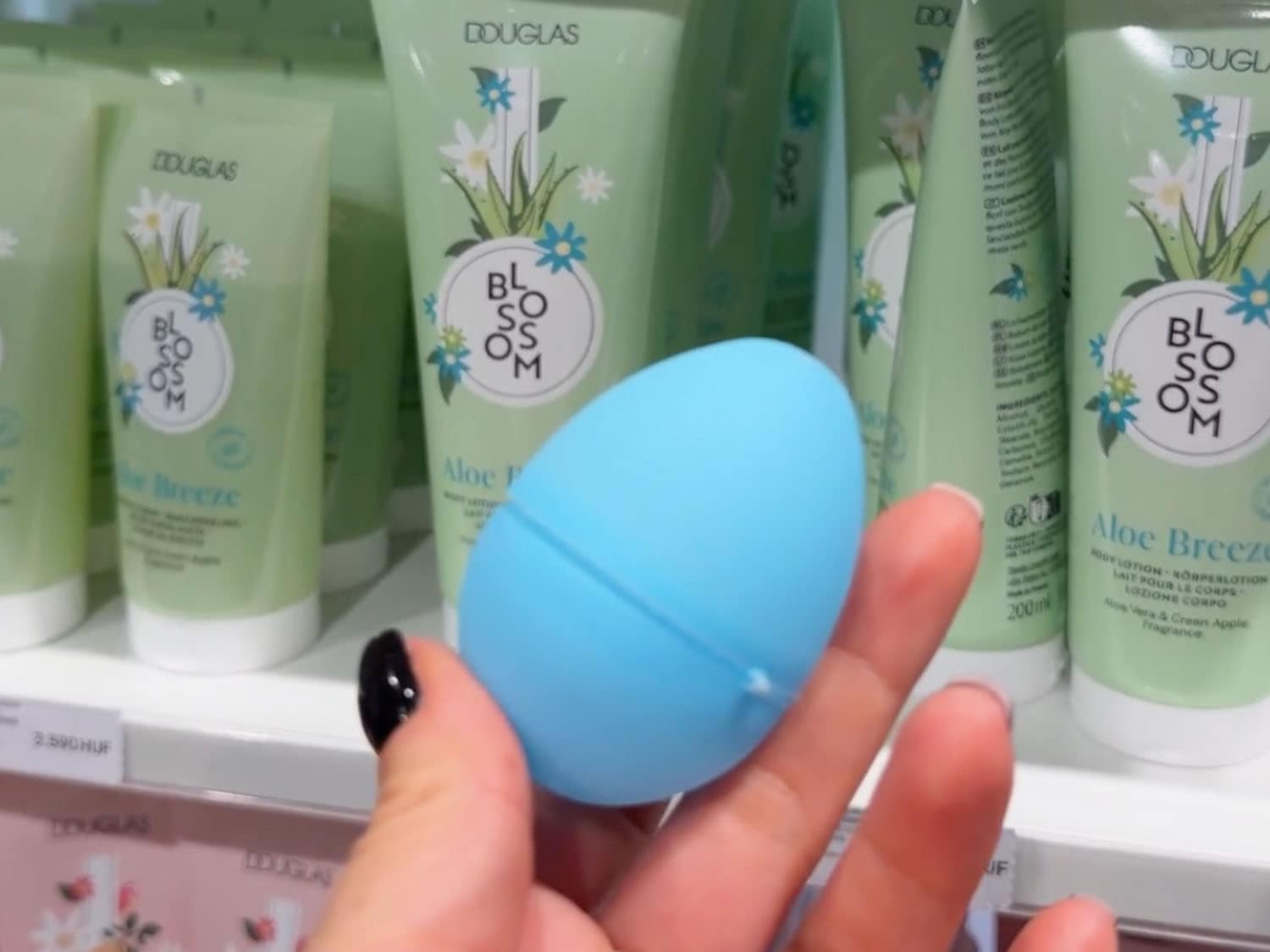 V popřední je modrá houbička na make-up ve tvaru vajíčka, v pozadí jsou zelené tuby tělového mléka Douglas Blossom Aloe Breeze s aloe vera a vůní zeleného jablka.