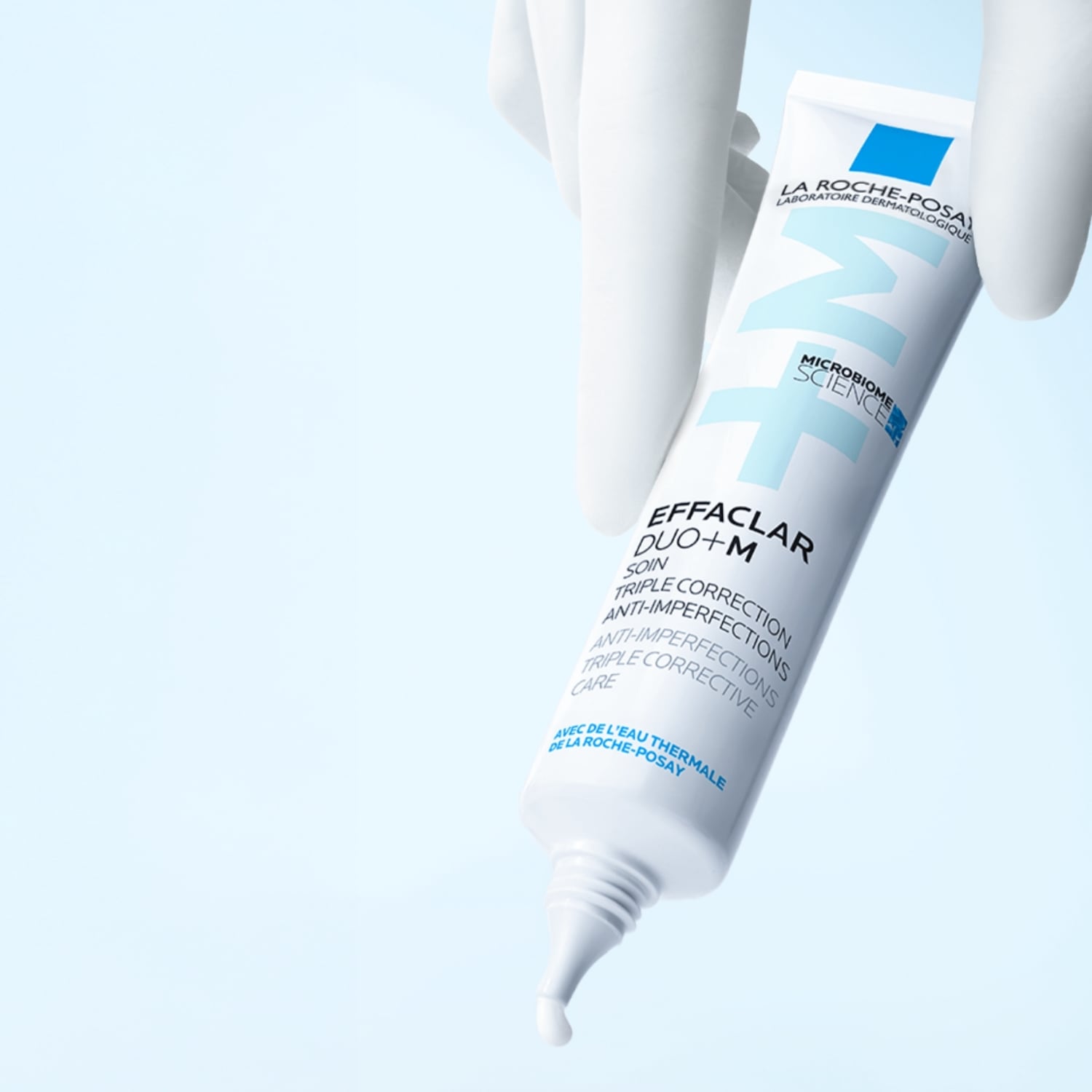 Gant blanc tenant un tube blanc de La Roche-Posay Effaclar Duo+M, soin triple correction anti-imperfections, avec une goutte de crème blanche qui en sort.
