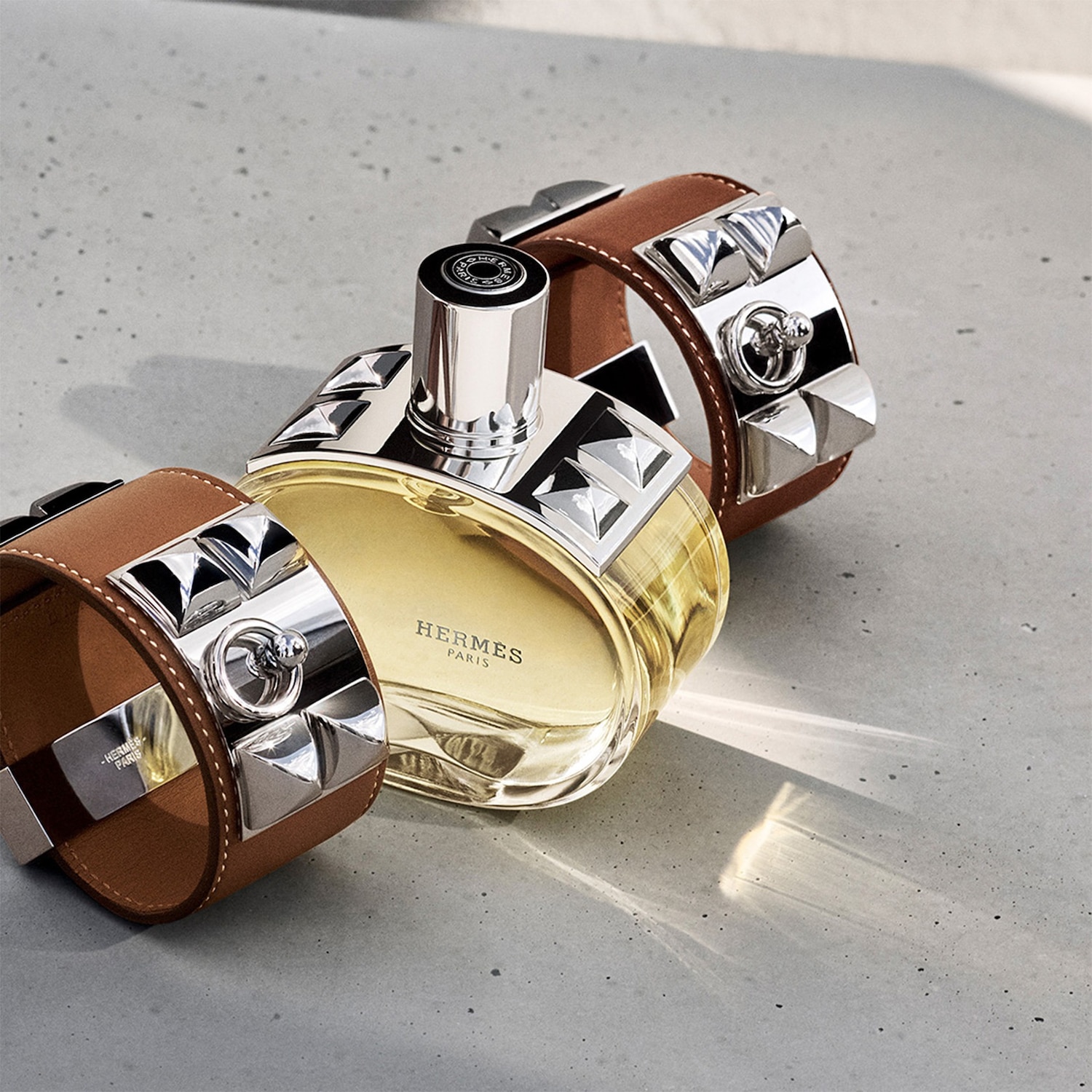 Een Hermès parfumflesje met goudkleurige vloeistof en zilverkleurige dop ligt tussen twee bruine leren armbanden met zilveren studs en ringen op een grijze ondergrond.