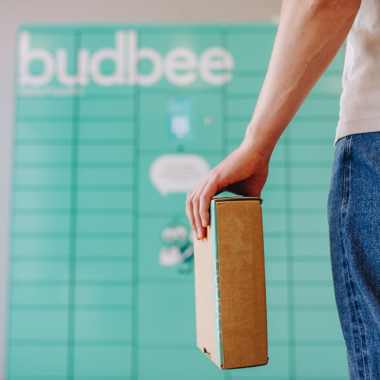 Een persoon houdt een bruine doos vast voor een turquoise Budbee-kluis, klaar om een pakket veilig te verzenden of op te halen.
