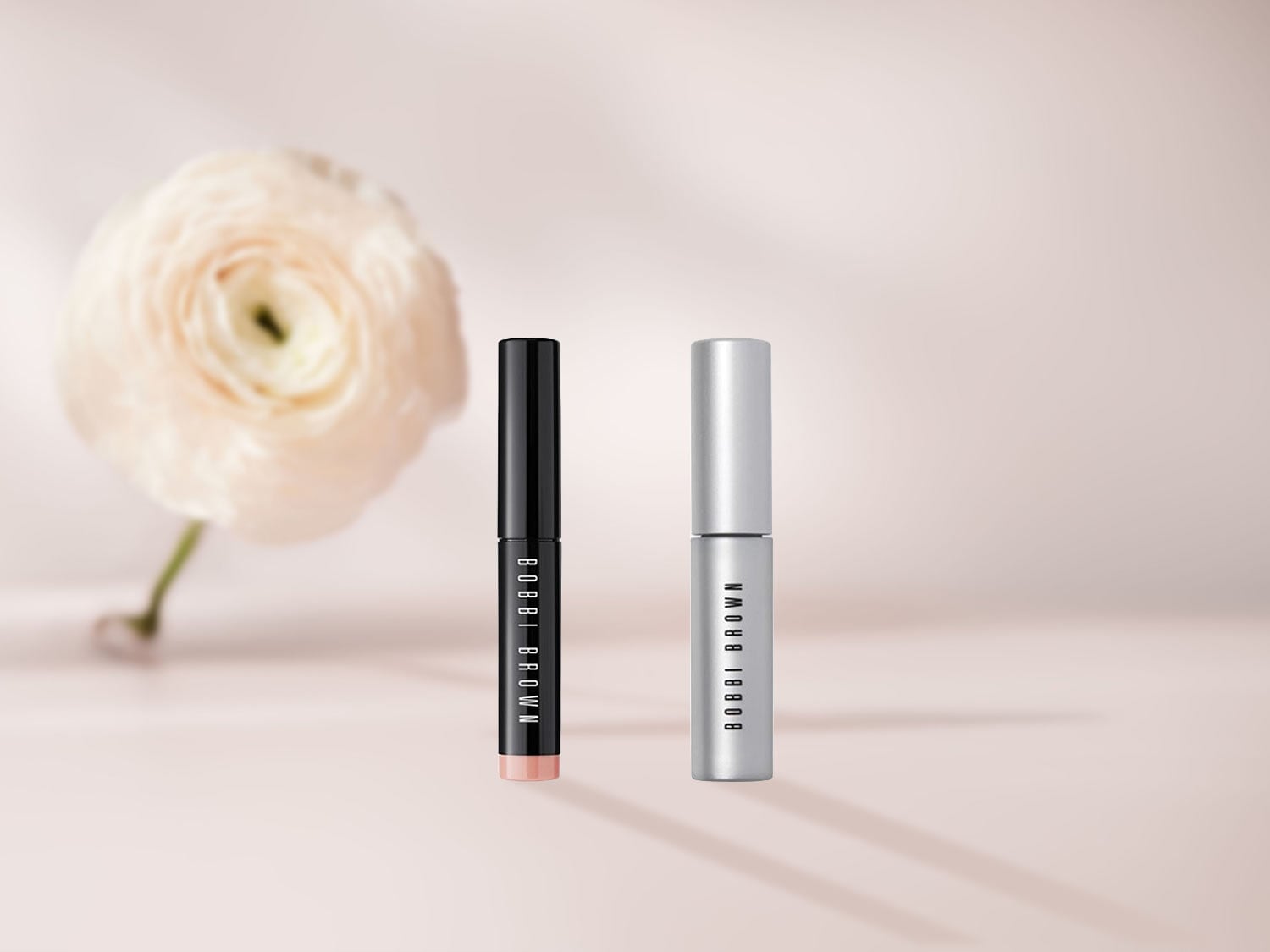 Zwei Bobbi Brown Lippenstifte, einer in Schwarz und einer in Silber, stehen vor einer zarten Pfingstrose.