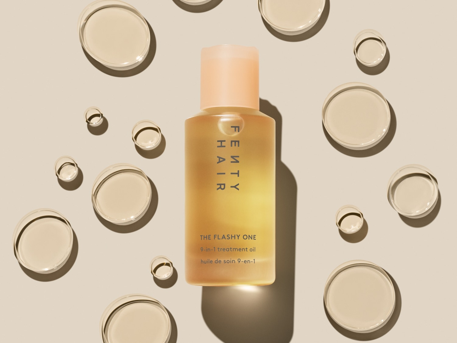 FENTY HAIR 'The Flashy One' 9-in-1 behandelingsolie in een heldere fles met een perzikkleurige dop, omringd door oliedruppels.
