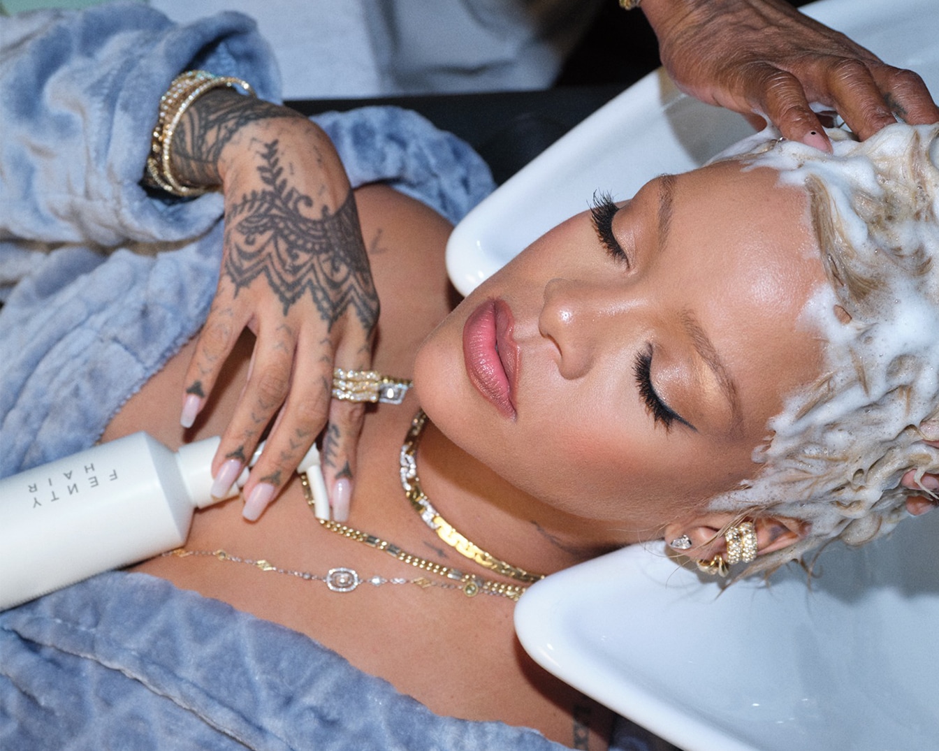 Rihanna ligt ontspannen met haar ogen dicht terwijl haar haar gewassen wordt met Fenty Hair producten, gekleed in een blauwe badjas en versierd met gouden sieraden.