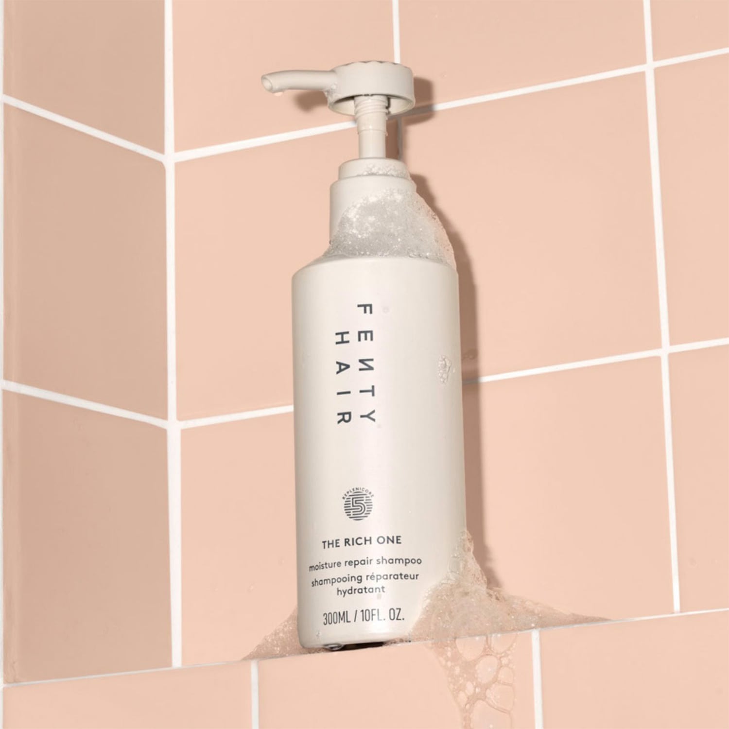 De Fenty Hair The Rich One shampoo in een witte fles met pompdispenser staat in een roze betegelde douche en is bedekt met schuim.