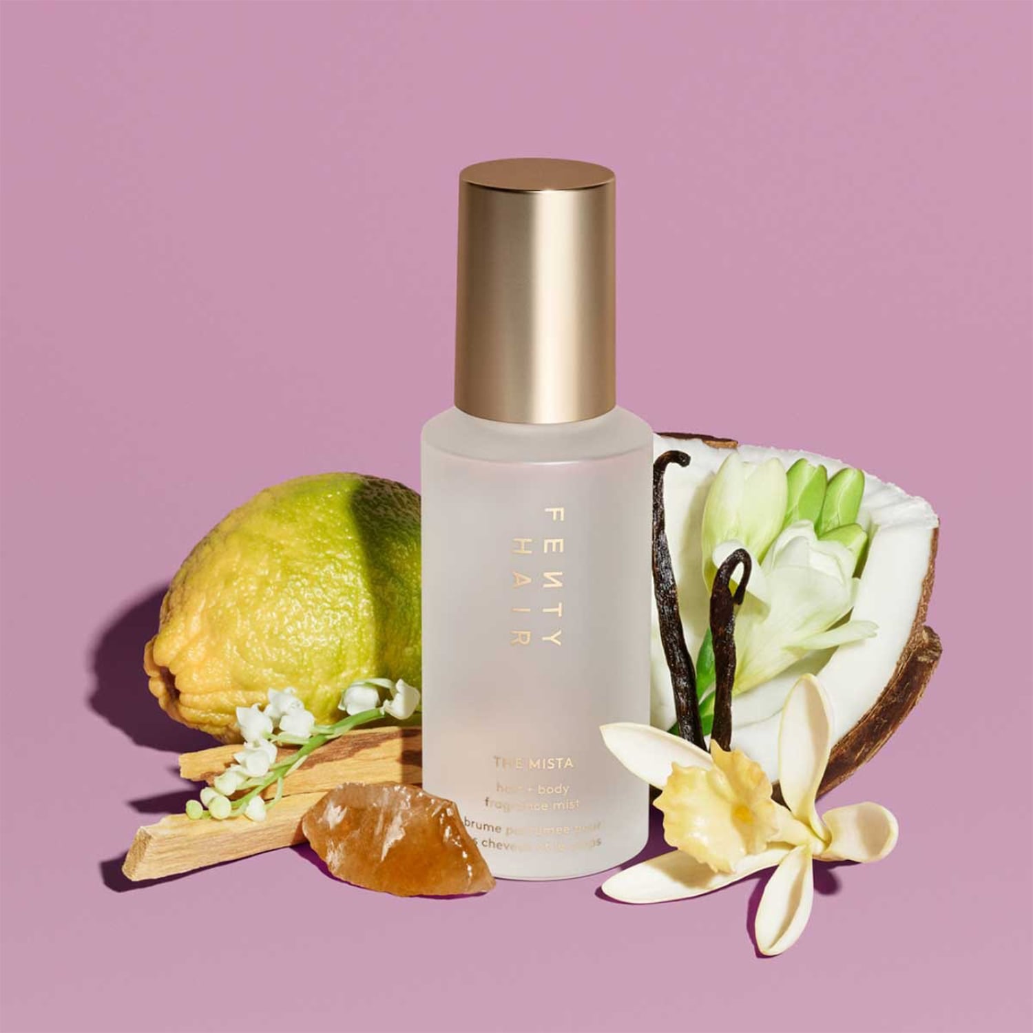 Fenty Hair The Mista haar- en bodymist in een heldere fles met gouden dop, omringd door citroen, kokosnoot, vanille en bloemen tegen een paarse achtergrond.