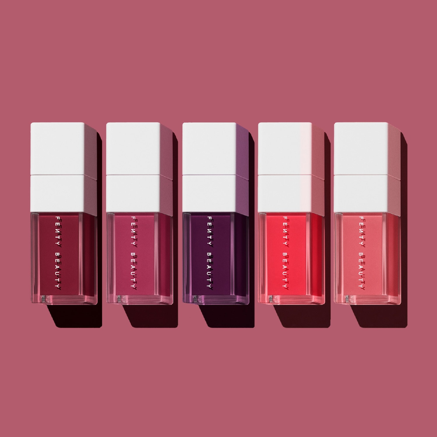 Vijf Fenty Beauty lipsticks in verschillende tinten rood en paars, elk met een vierkante witte dop, staan op een roze achtergrond.