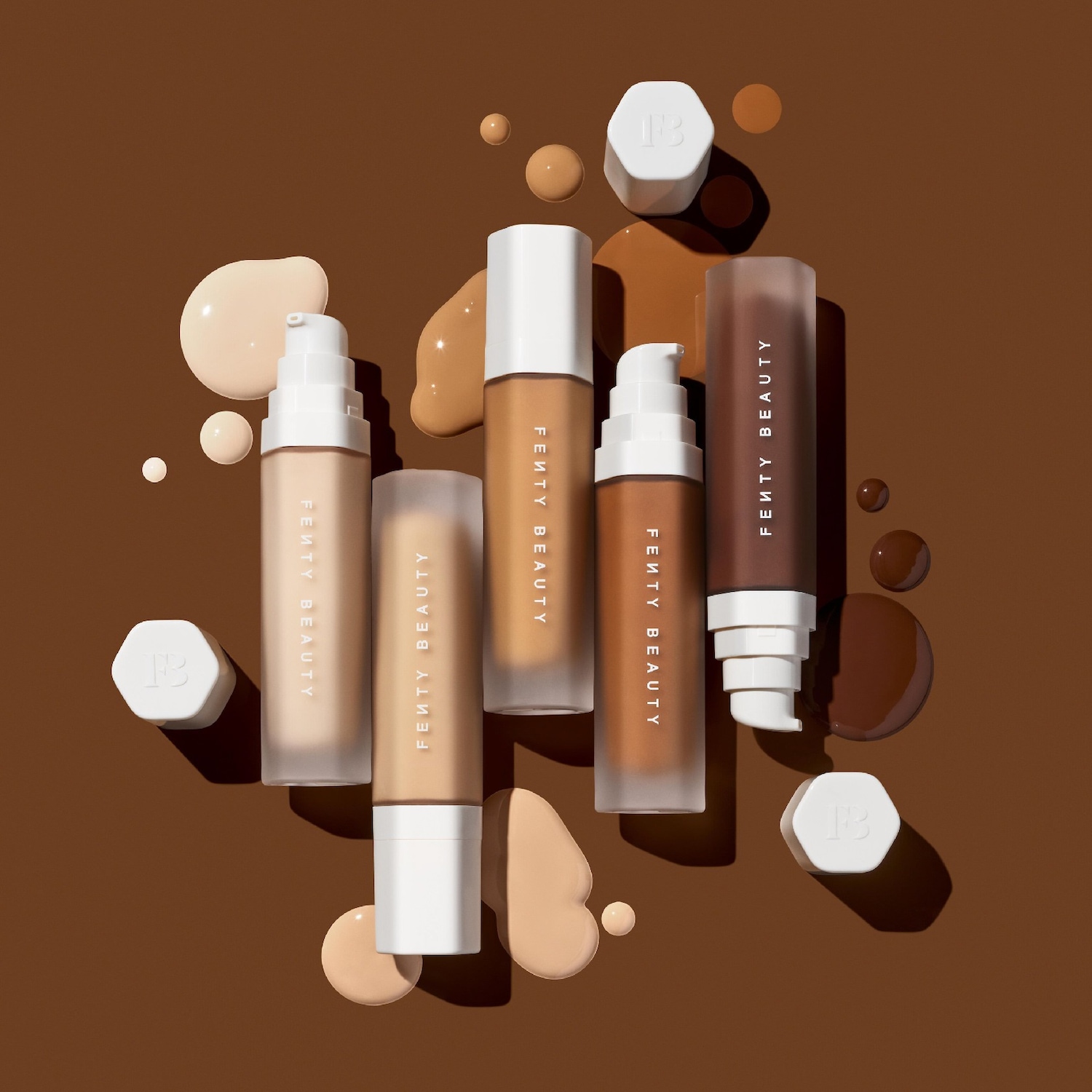 Verschillende tinten Fenty Beauty foundation in flessen met witte doppen, omringd door foundationdruppels op een bruine achtergrond.