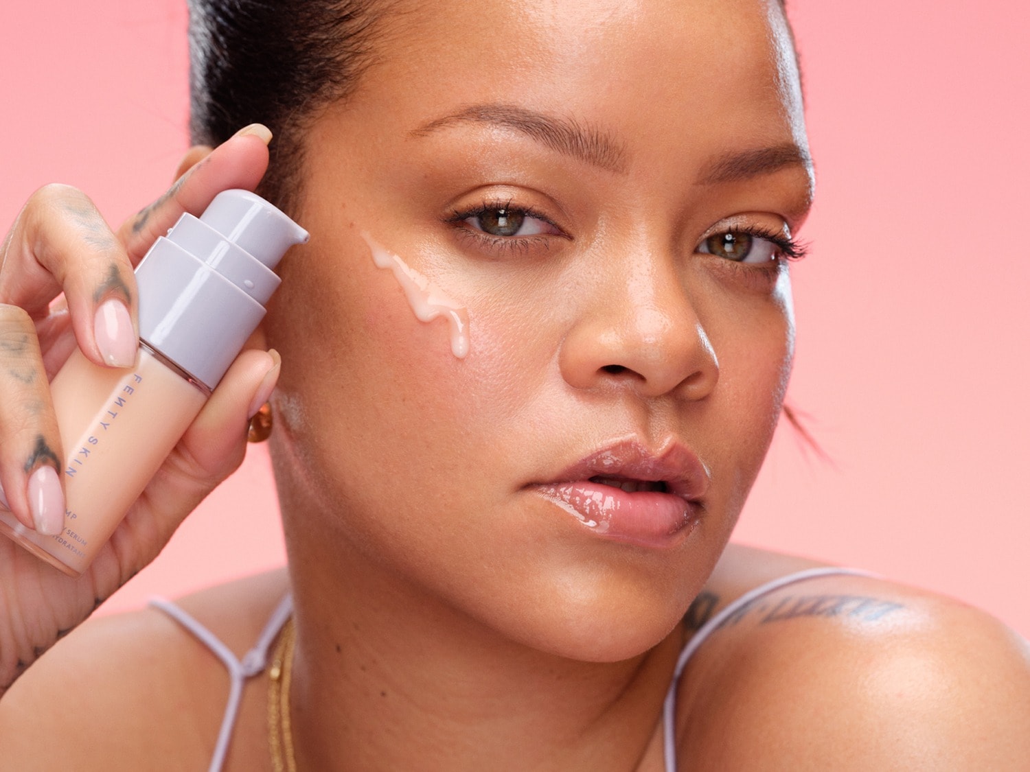 Een vrouw met een stralende huid brengt Fenty Skin hydraterend serum aan, verpakt in een zachtbeige fles met een lichtgrijze pomp.