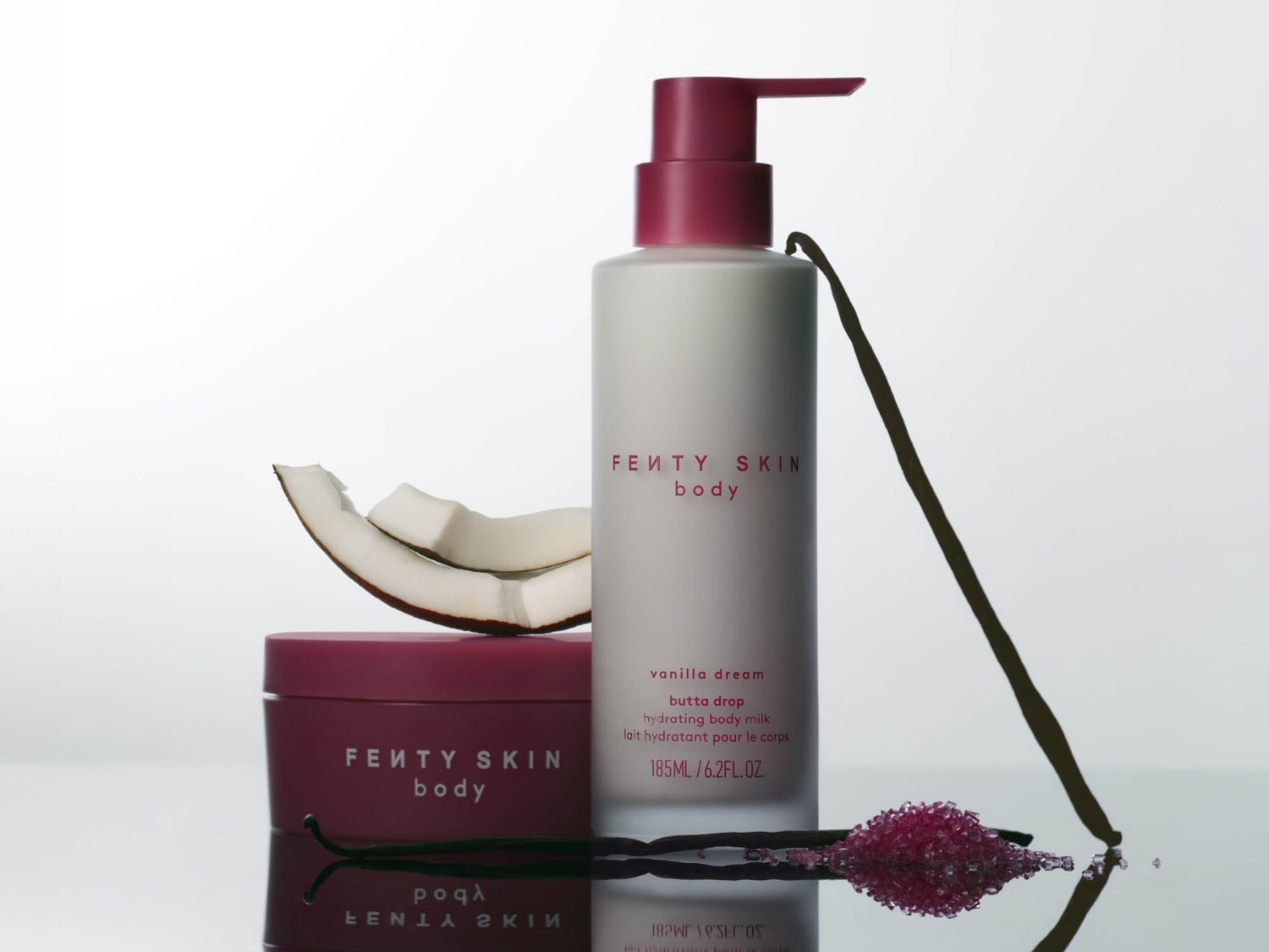 Ontdek Fenty Skin Body's Butta Drop hydraterende bodymilk in een witte fles met een roze pomp, naast een pot en kokosnoot, voor een heerlijke vanille droom.