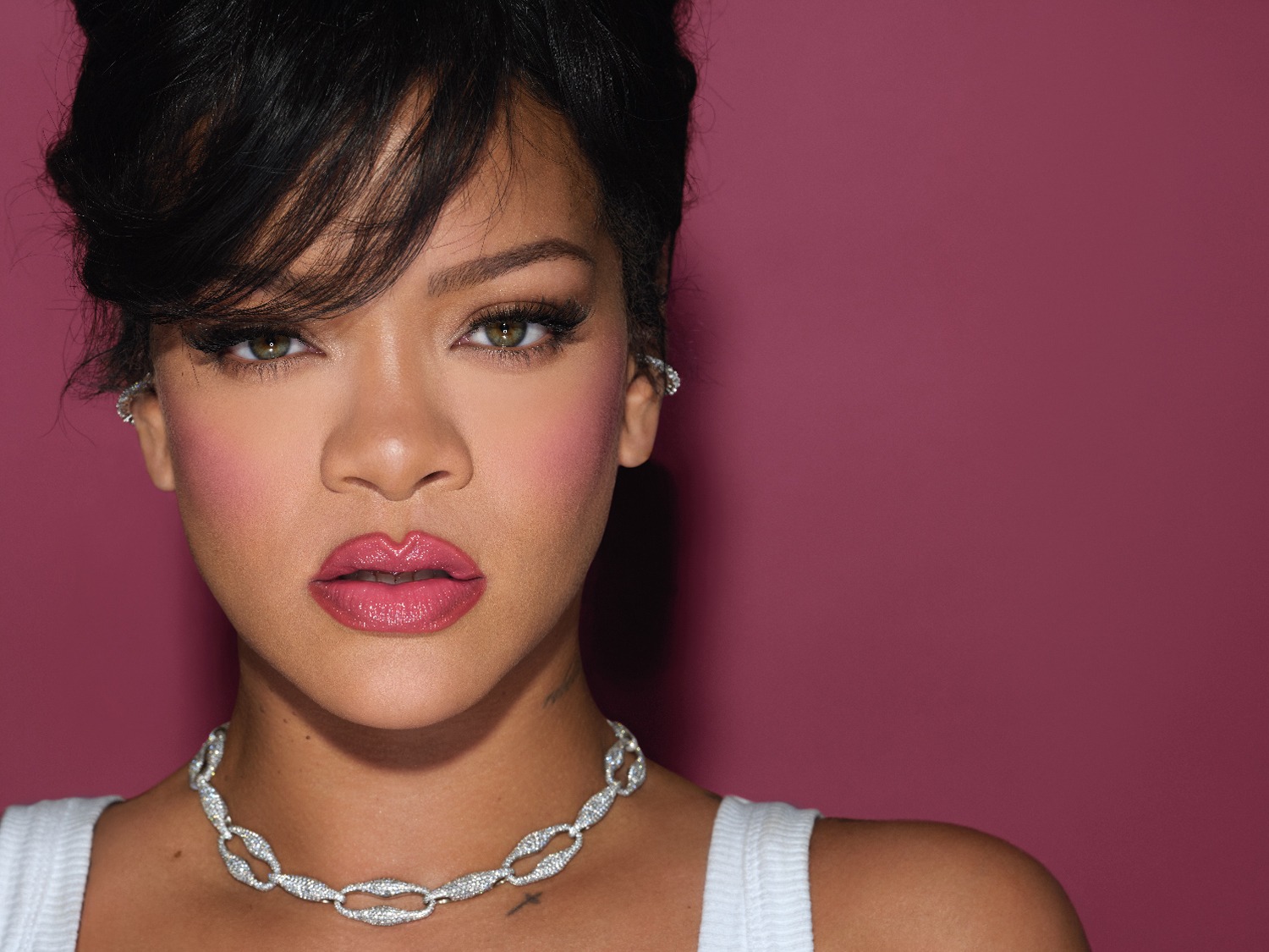 Gros plan sur Rihanna avec un rouge à lèvres rose brillant, un fard à joues rose et un collier en argent étincelant sur un fond rose.