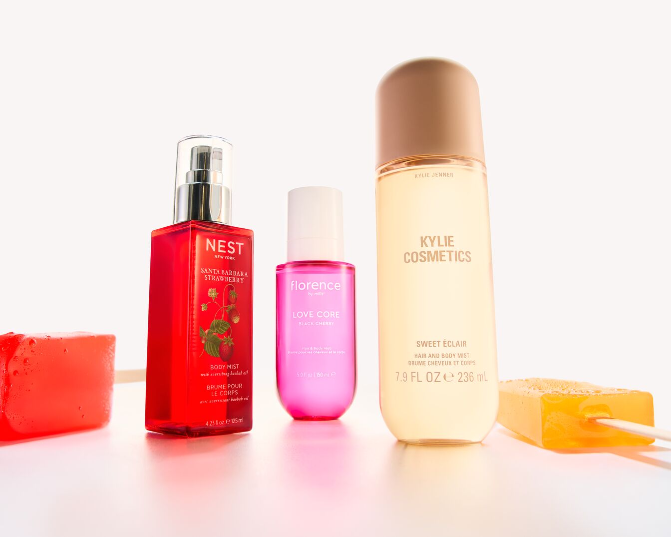 Trei sticle de spray de corp, una roșie de la Nest New York, una roz de la Florence by Mills și una galbenă de la Kylie Cosmetics, alături de două batoane de săpun colorate.