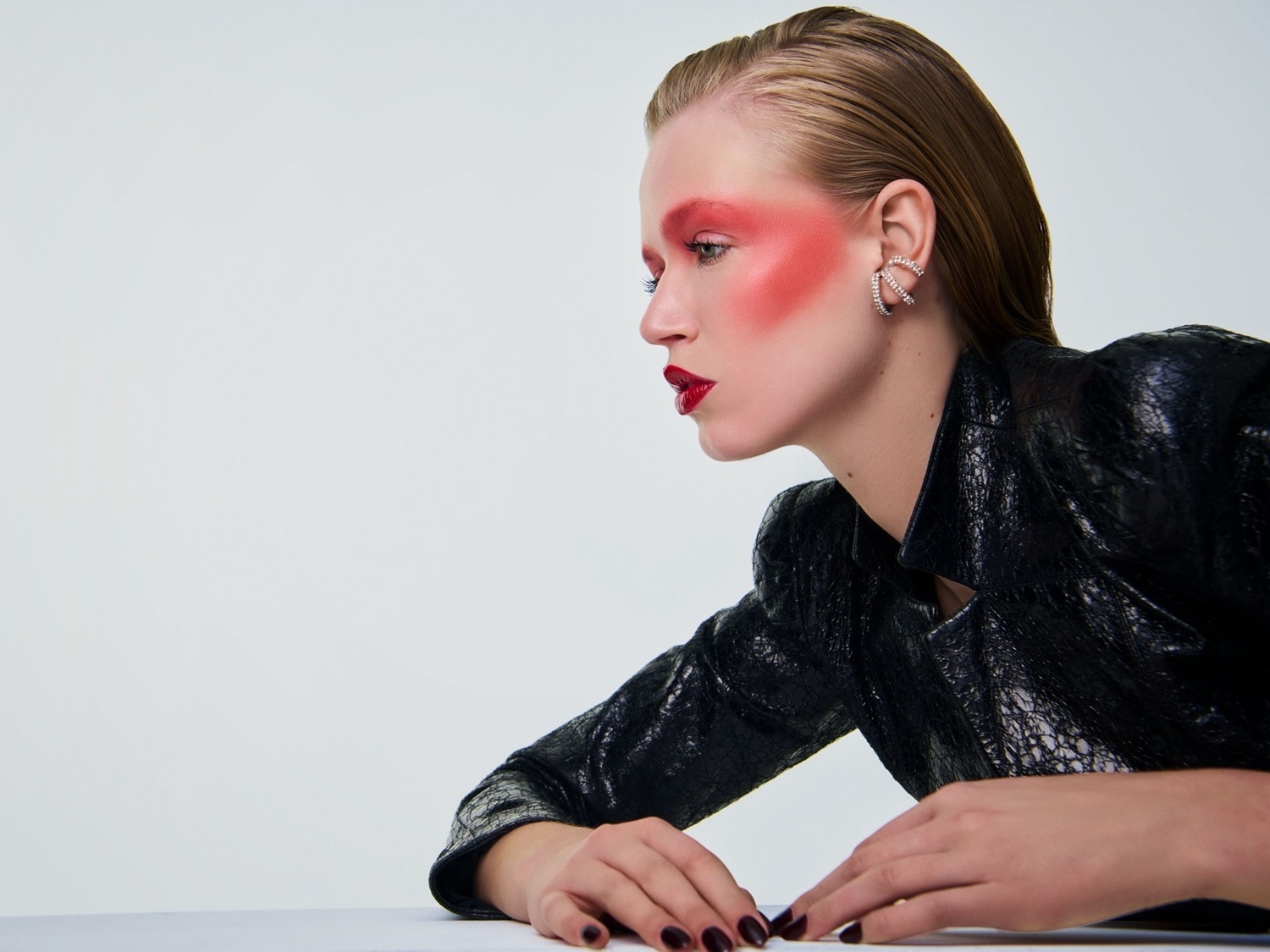 Profilansicht einer Frau mit rotem Rouge, roten Lippen und glänzendem Haar, die eine schwarze Lederjacke und Ohrringe trägt.