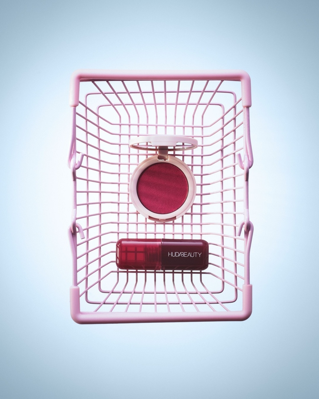 In einem rosa Drahtkorb liegen ein Rouge mit geöffnetem Deckel und ein Lippenstift von Huda Beauty in verschiedenen Rottönen.