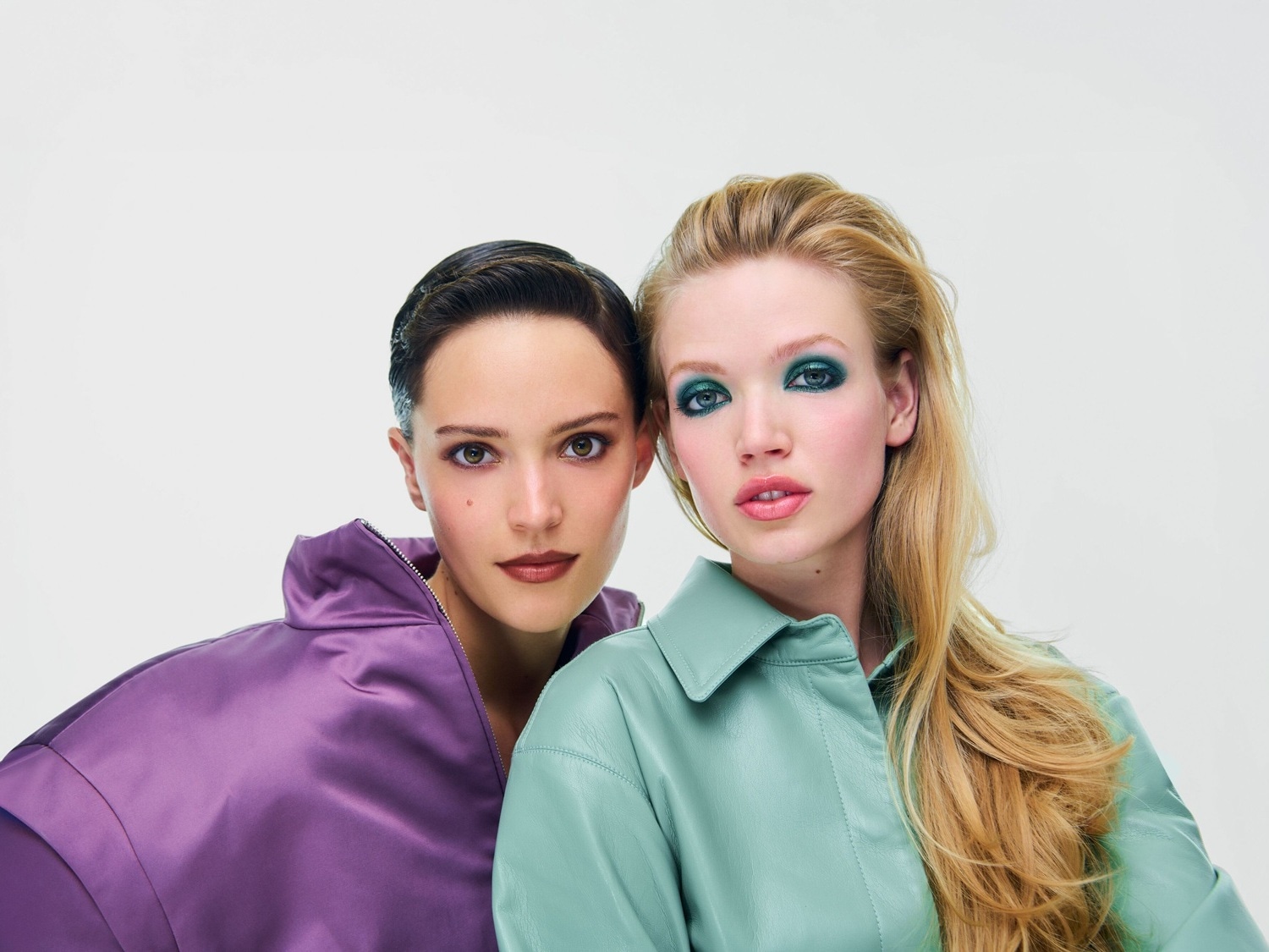 Zwei Models mit auffälligem grünen Lidschatten, die eine trägt eine lila Jacke, die andere eine hellgrüne Jacke, beide blicken in die Kamera.