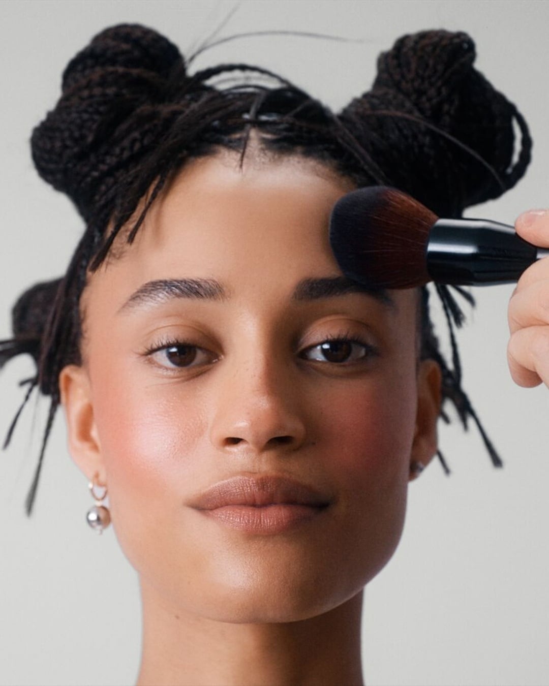 Une femme applique du blush sur sa joue avec un pinceau à maquillage, créant un look frais et éclatant.