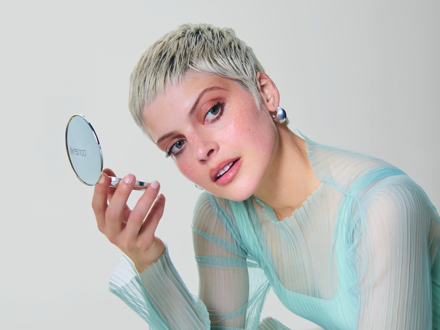 O femeie cu păr blond scurt și un top transparent albastru deschis ține în mână o oglindă compactă Douglas argintie și își privește reflexia.