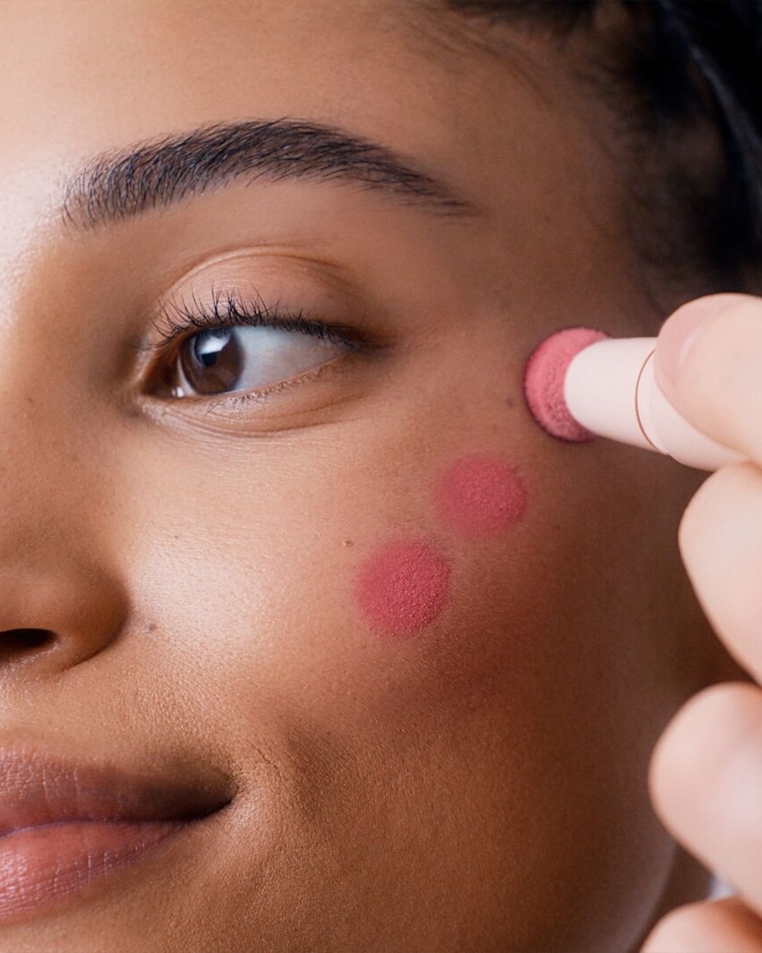 Gros plan d'une femme appliquant un blush rose crémeux sur sa joue avec un applicateur rond, pour un look frais et éclatant.