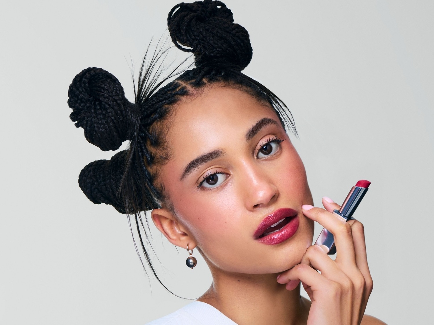 Une femme avec des tresses en chignon applique un rouge à lèvres rouge foncé brillant, rehaussant son maquillage rosé et ses boucles d'oreilles pendantes.