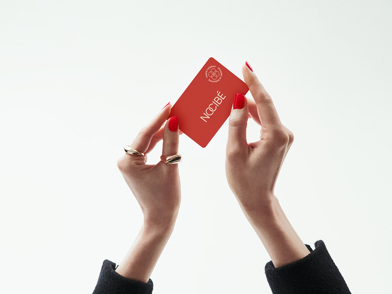 Les mains tiennent une carte cadeau rouge Nocibé, parfait pour offrir un moment de beauté et de bien-être.