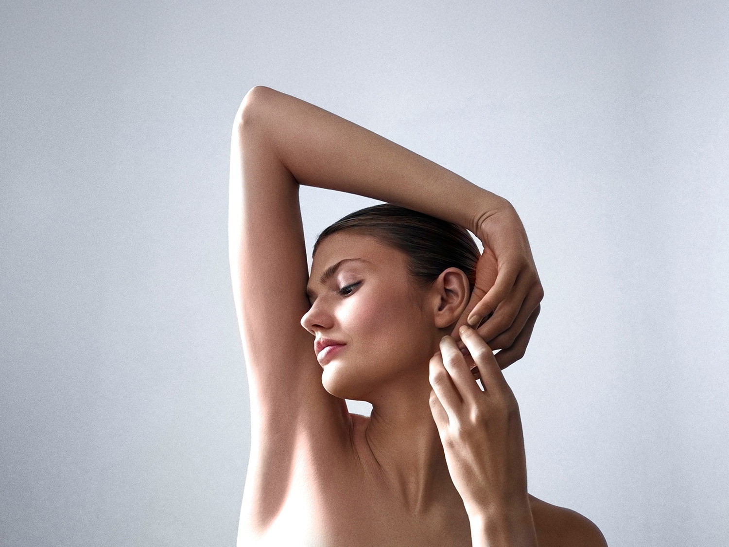 Une femme avec le bras levé, touchant son oreille, peau lisse et maquillage subtil, sur un fond gris clair.